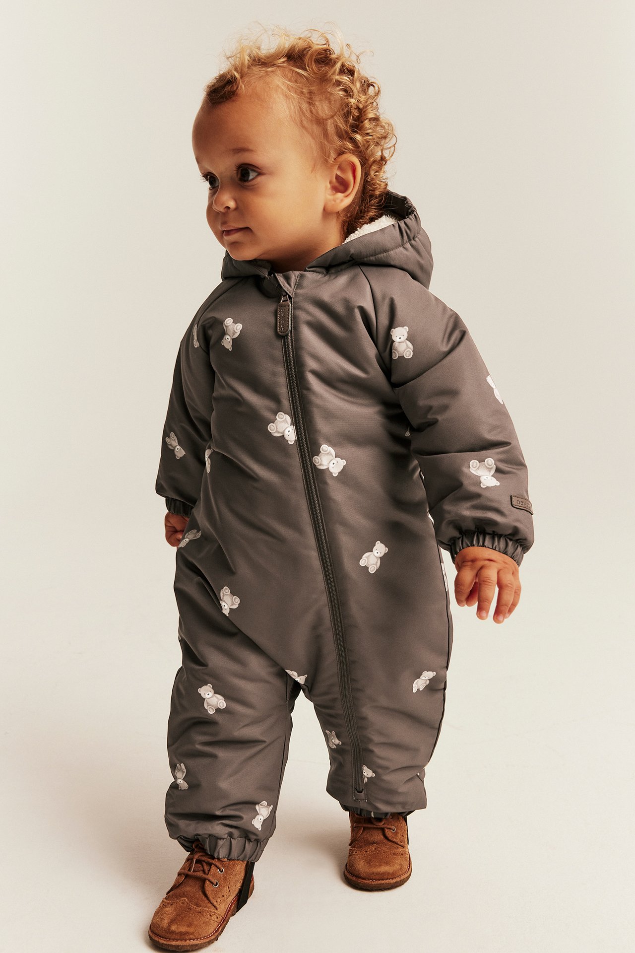 Front view: baby wears dark gray padded hooded jumpsuit with white teddy bear print and brown boots.