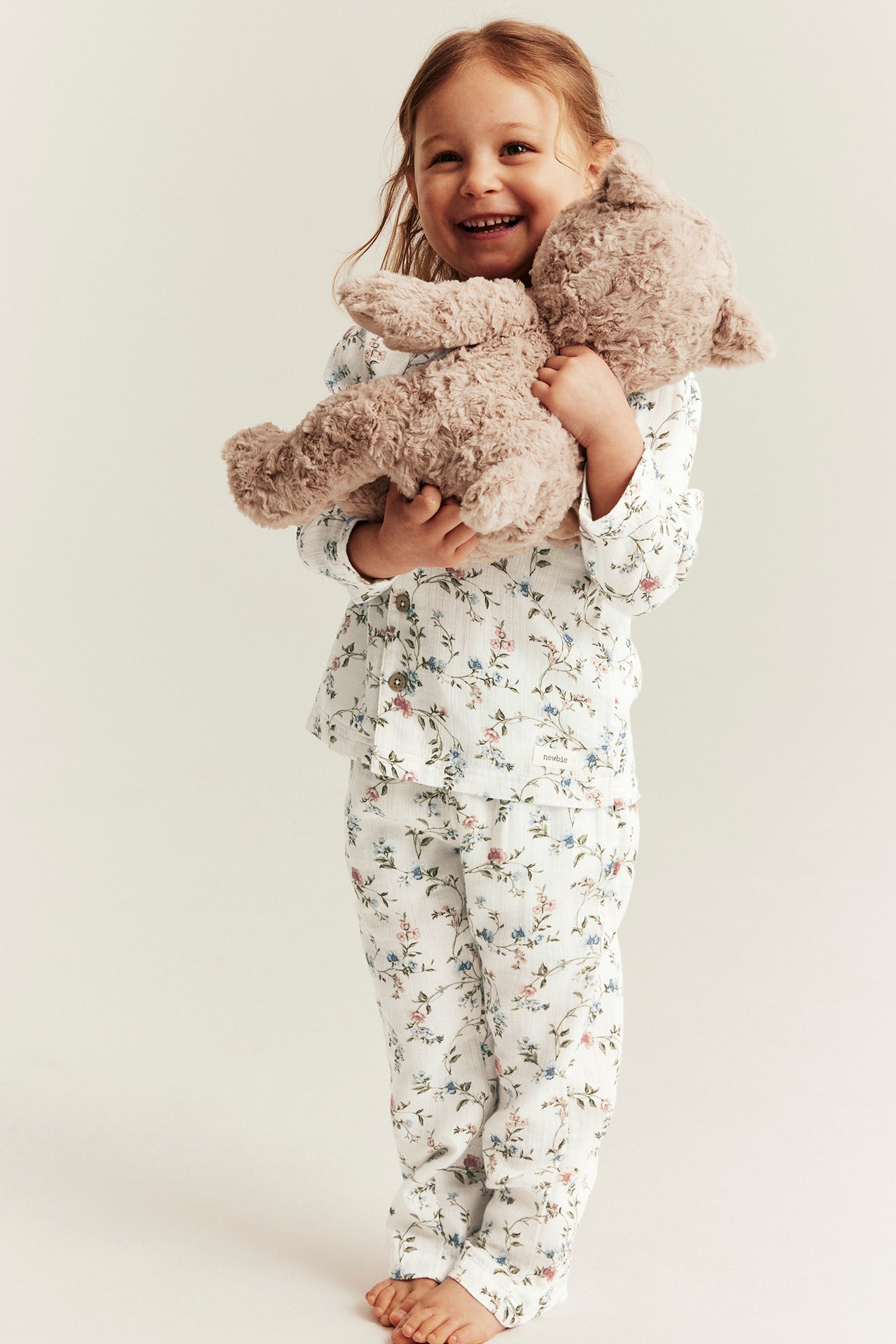 Front view: Smiling kid wears an off-white floral pyjama set with button-up top, holding a brown teddy bear.