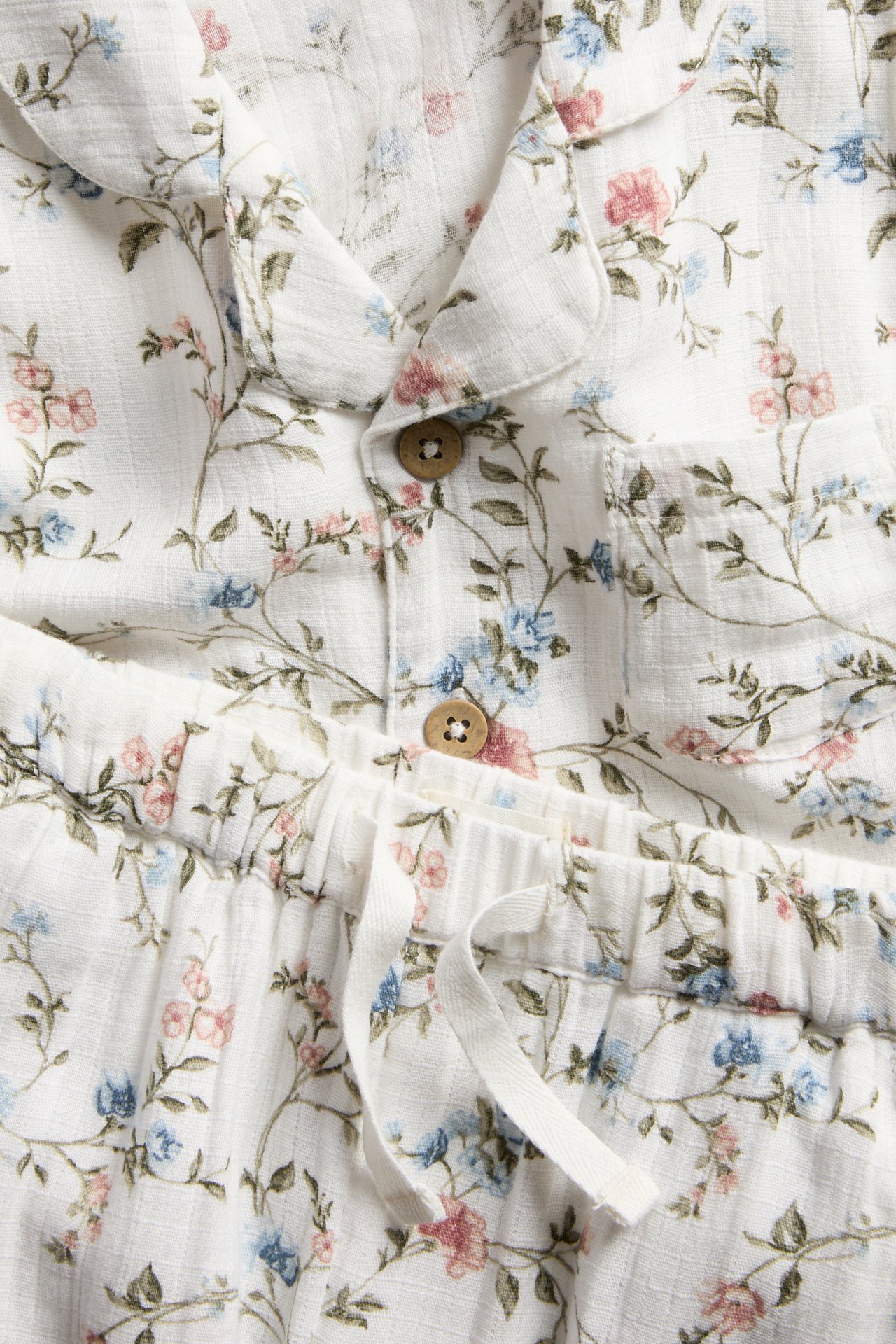 Close-up of offwhite cotton pyjamas with blue and pink floral pattern, wooden buttons, and a drawstring waist.