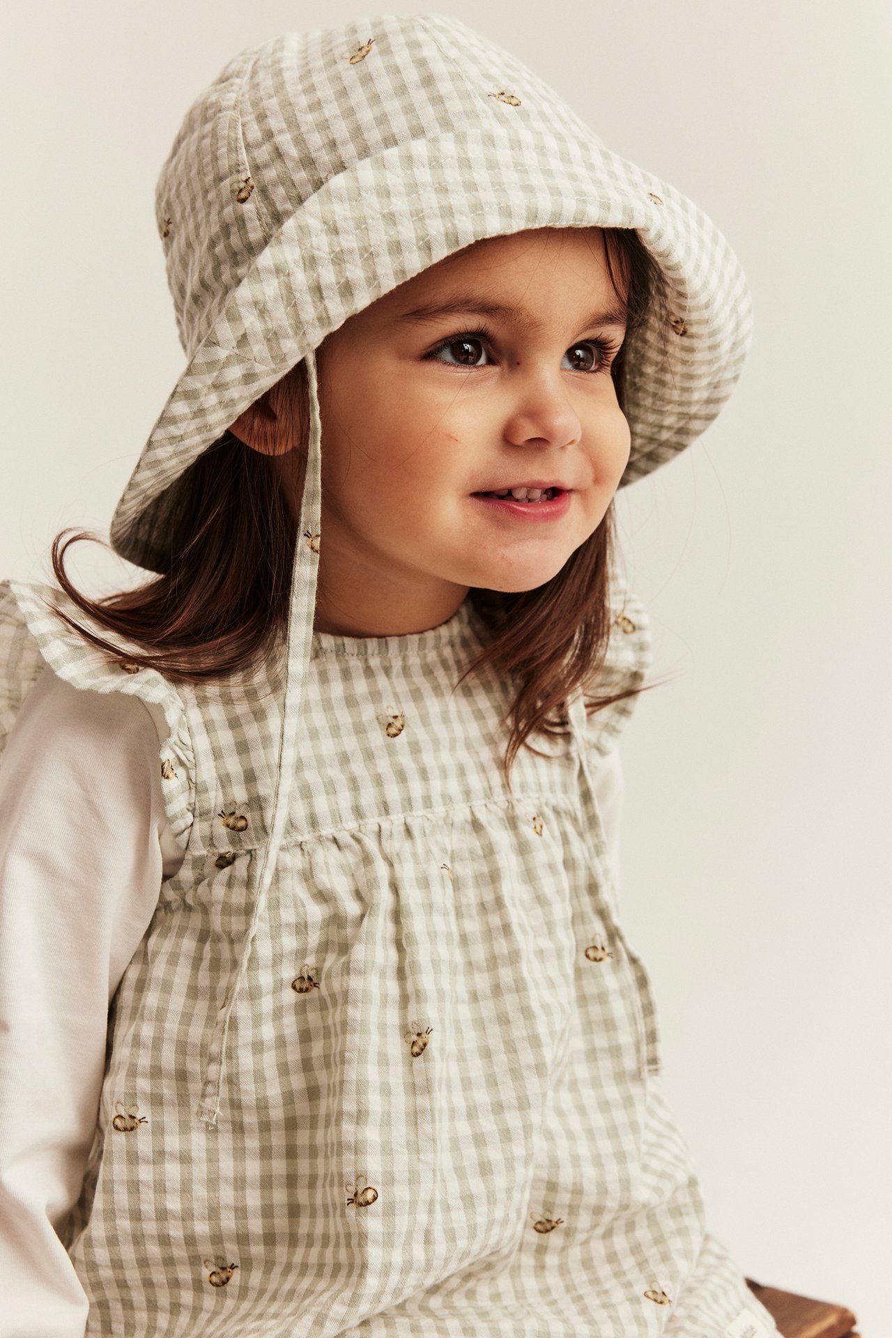 Close-up of a kid wearing a light green checked sun hat with bee embroidery, over a matching dress.
