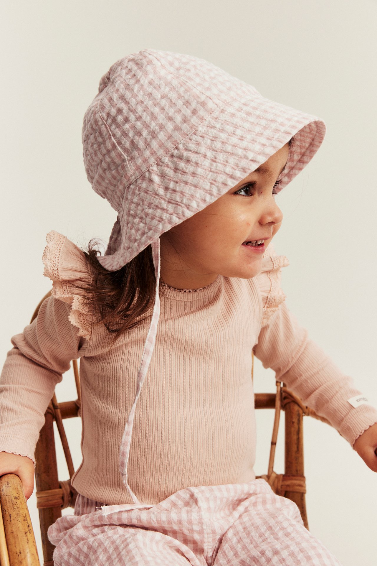 Side view: kid in light pink checked woven sun hat with ties, ribbed top and matching pants.