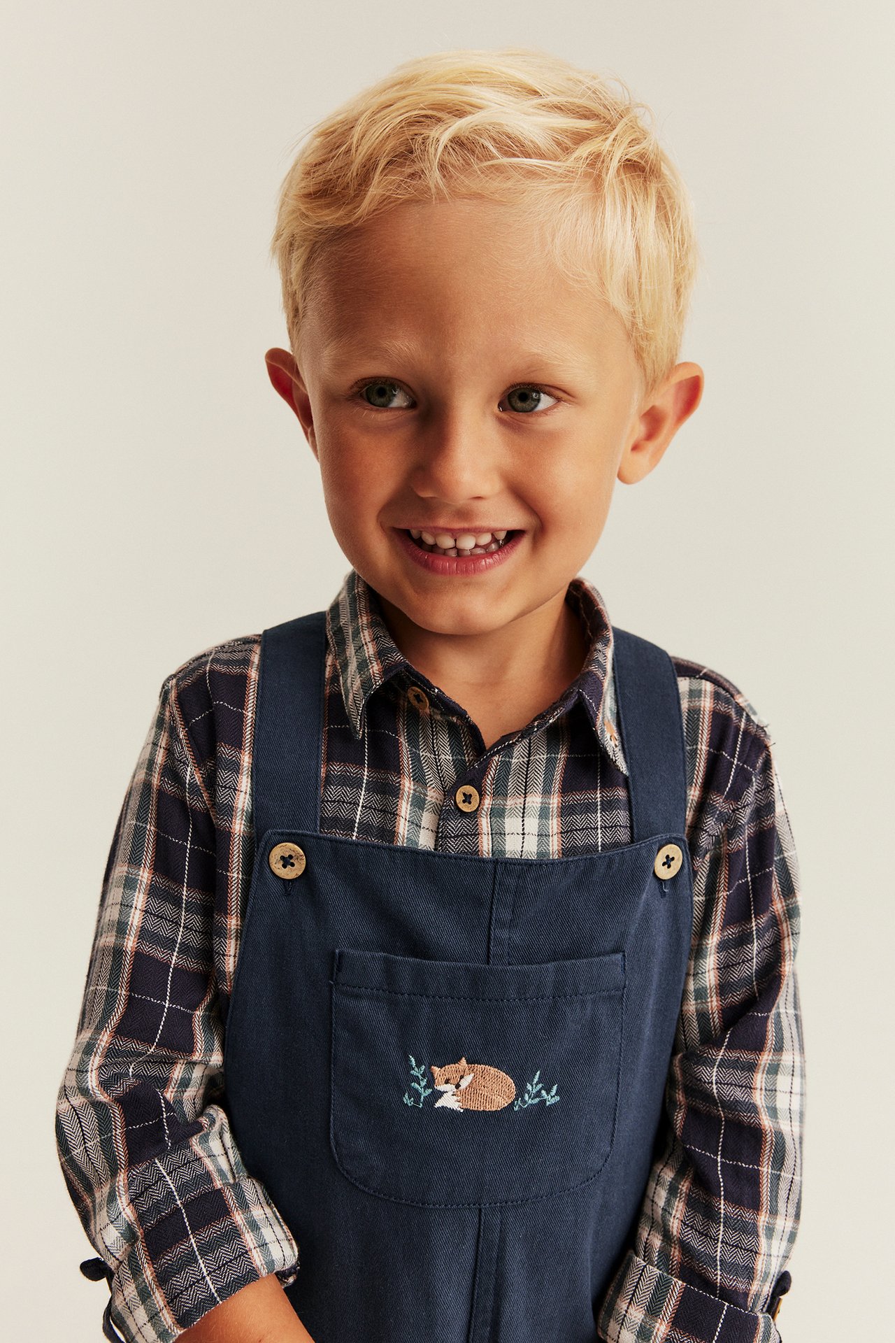Front view of a kid wearing a navy blue overall with a fox embroidery over a plaid shirt.