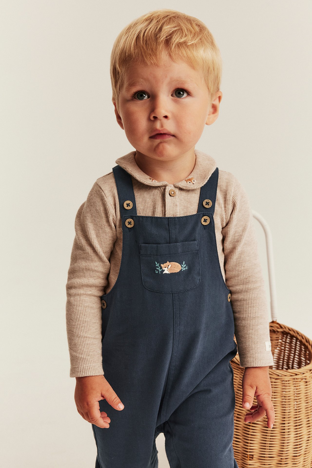 Front view of baby boy in dark blue overalls with fox embroidery over a beige long-sleeved ribbed top.