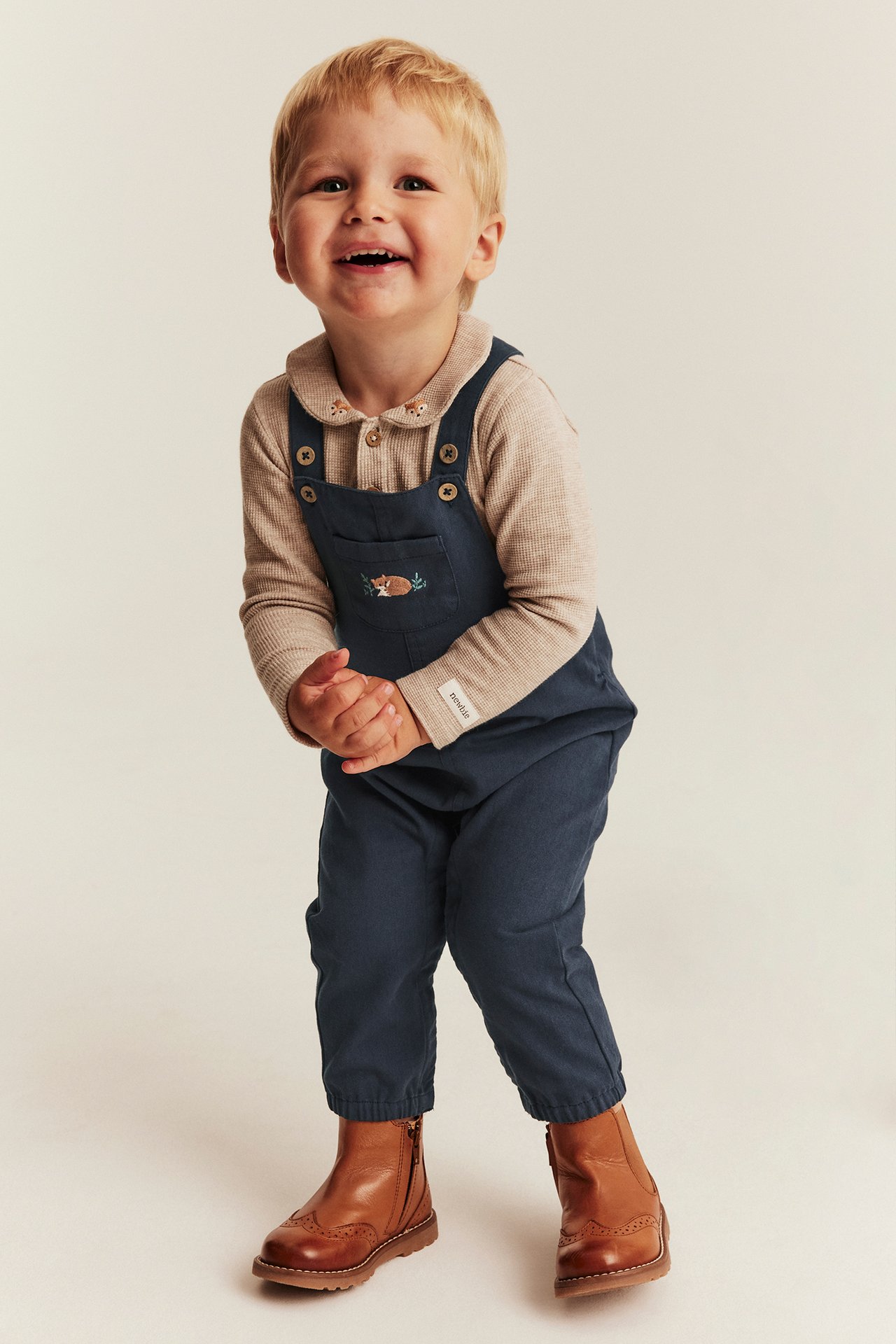 Front view of a kid wearing dark blue fox-embroidered overalls, a beige collared shirt, and brown boots.