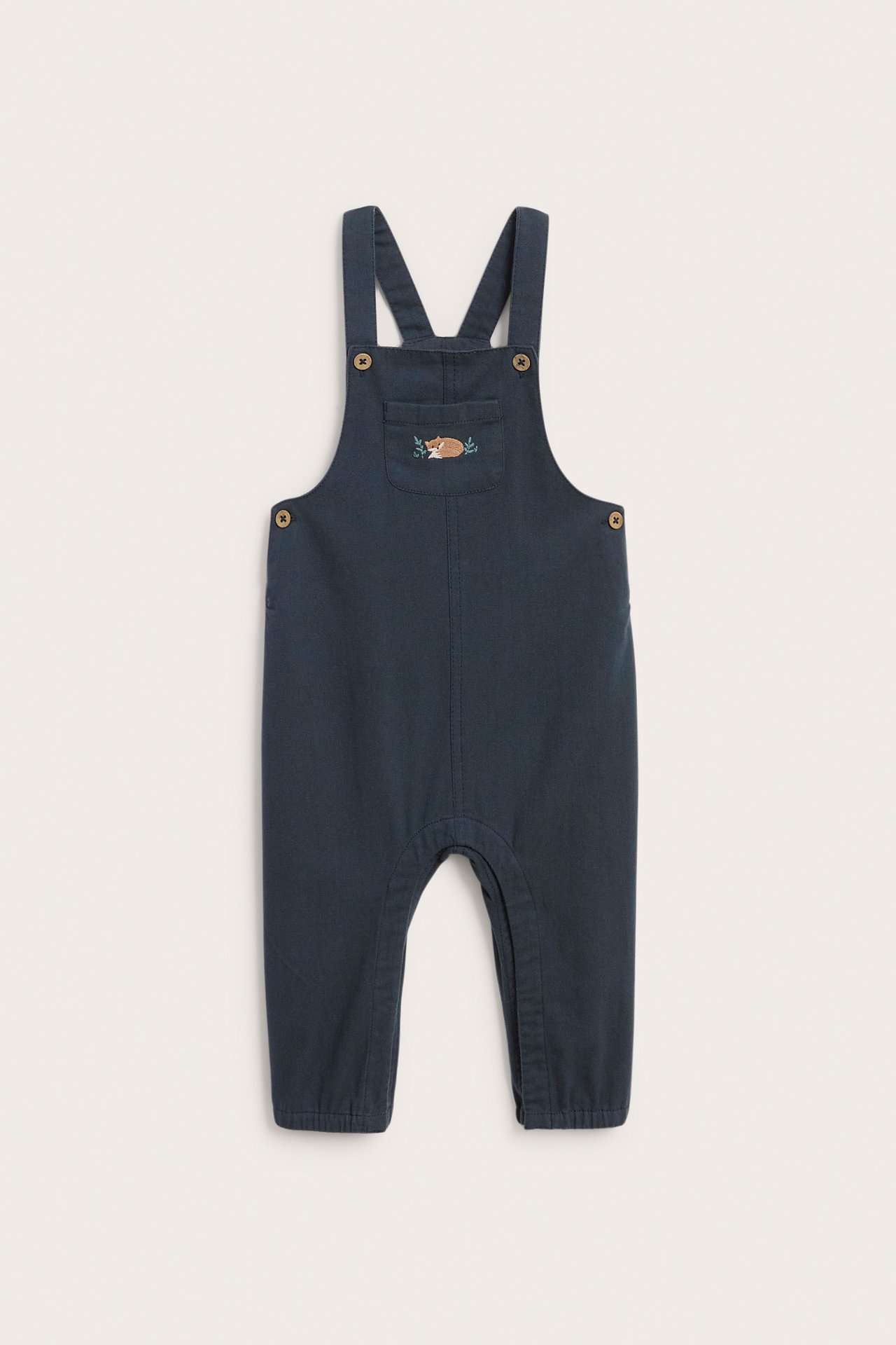 Front view of a dark blue baby overall with an embroidered sleeping fox and leaves on the chest pocket.