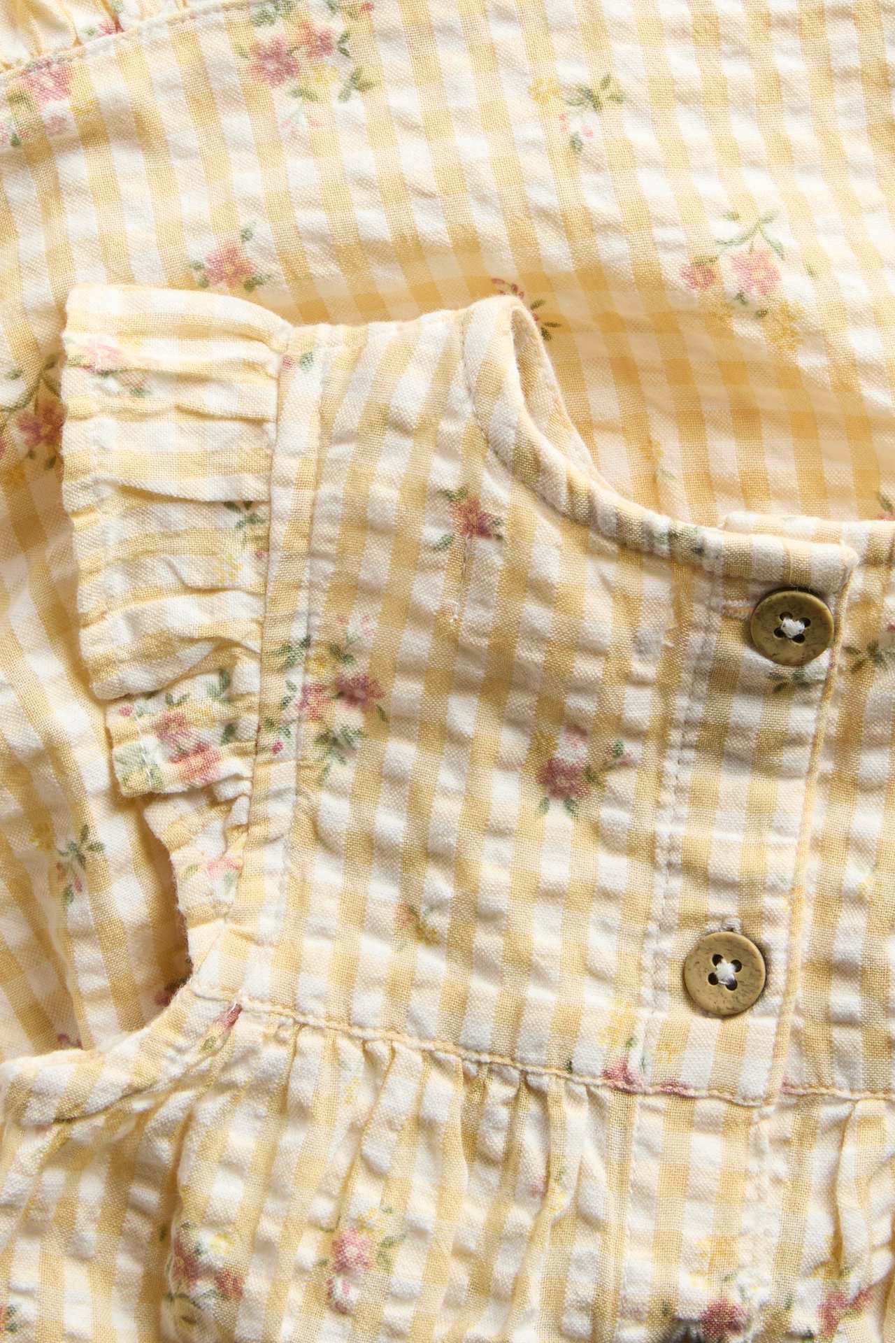 Close-up front view of a yellow and white gingham cotton dress with small floral print, ruffled sleeves, and brown buttons.