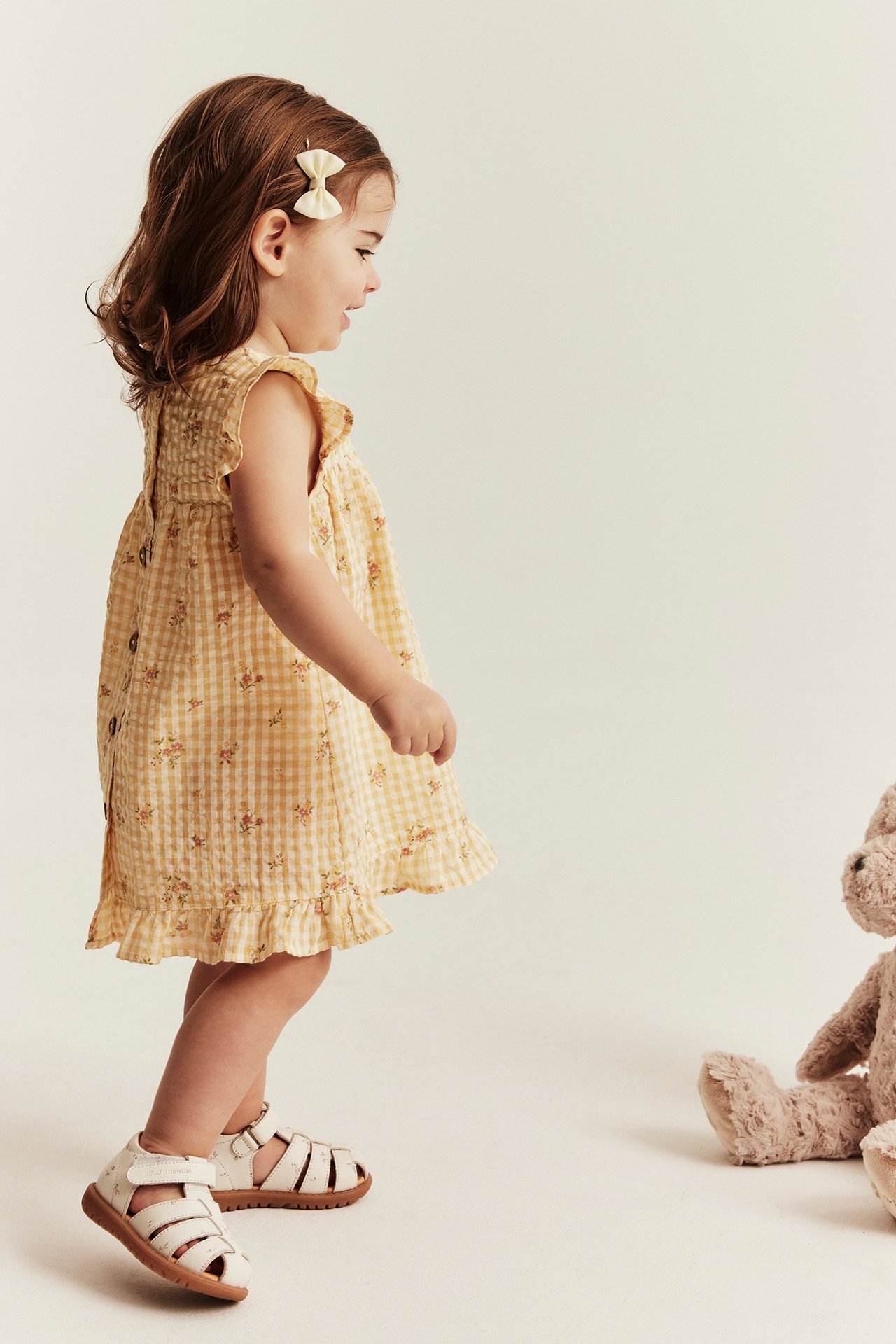 Side view of a kid in a yellow floral gingham dress with ruffled shoulders, white sandals, bow clip.