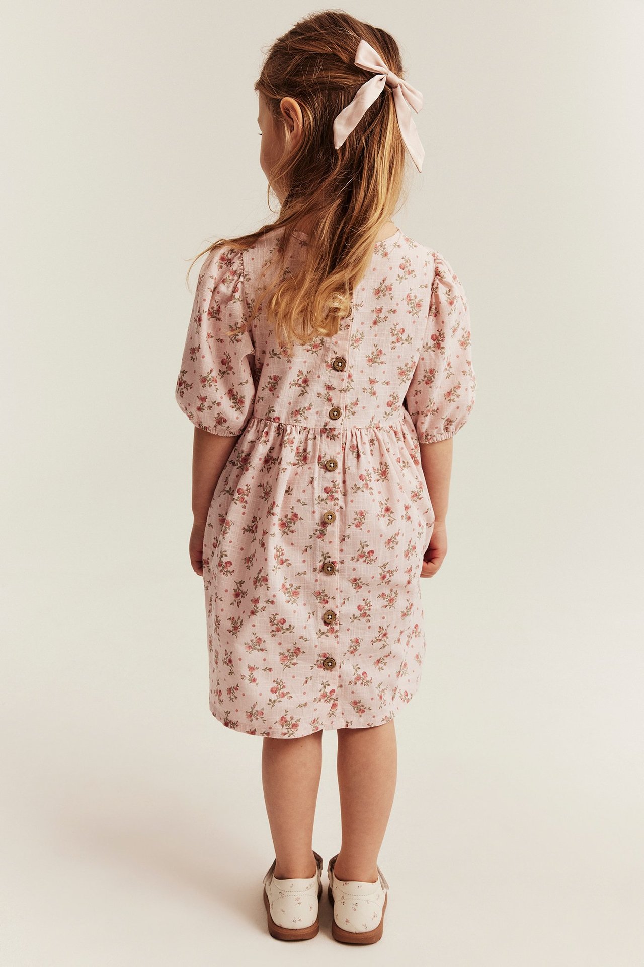 Back view: kid in a pink floral puff-sleeve dress with back buttons, a pink hair bow, and shoes.