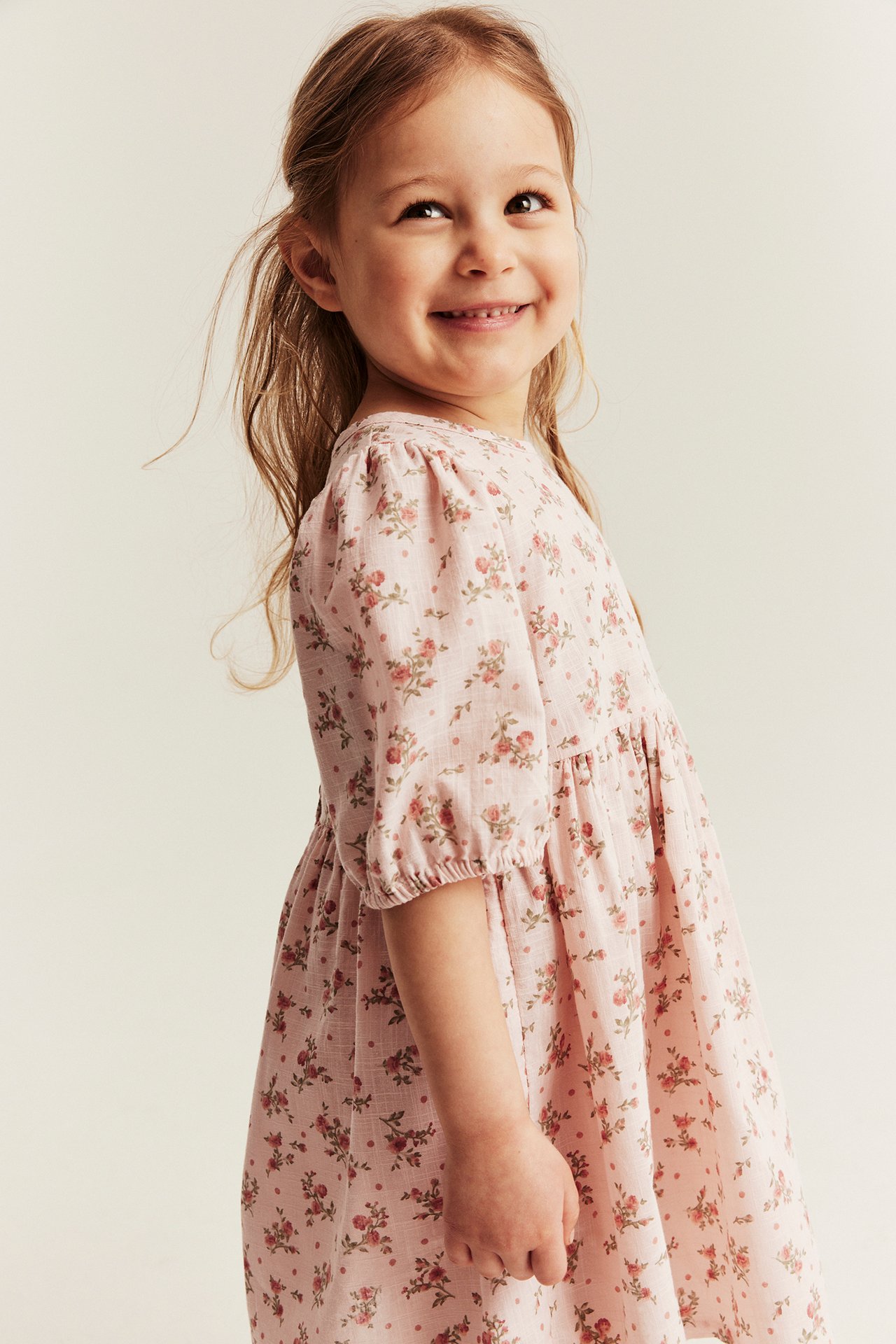 Side view of a smiling kid wearing a light pink floral dress with puff sleeves and elastic cuffs.