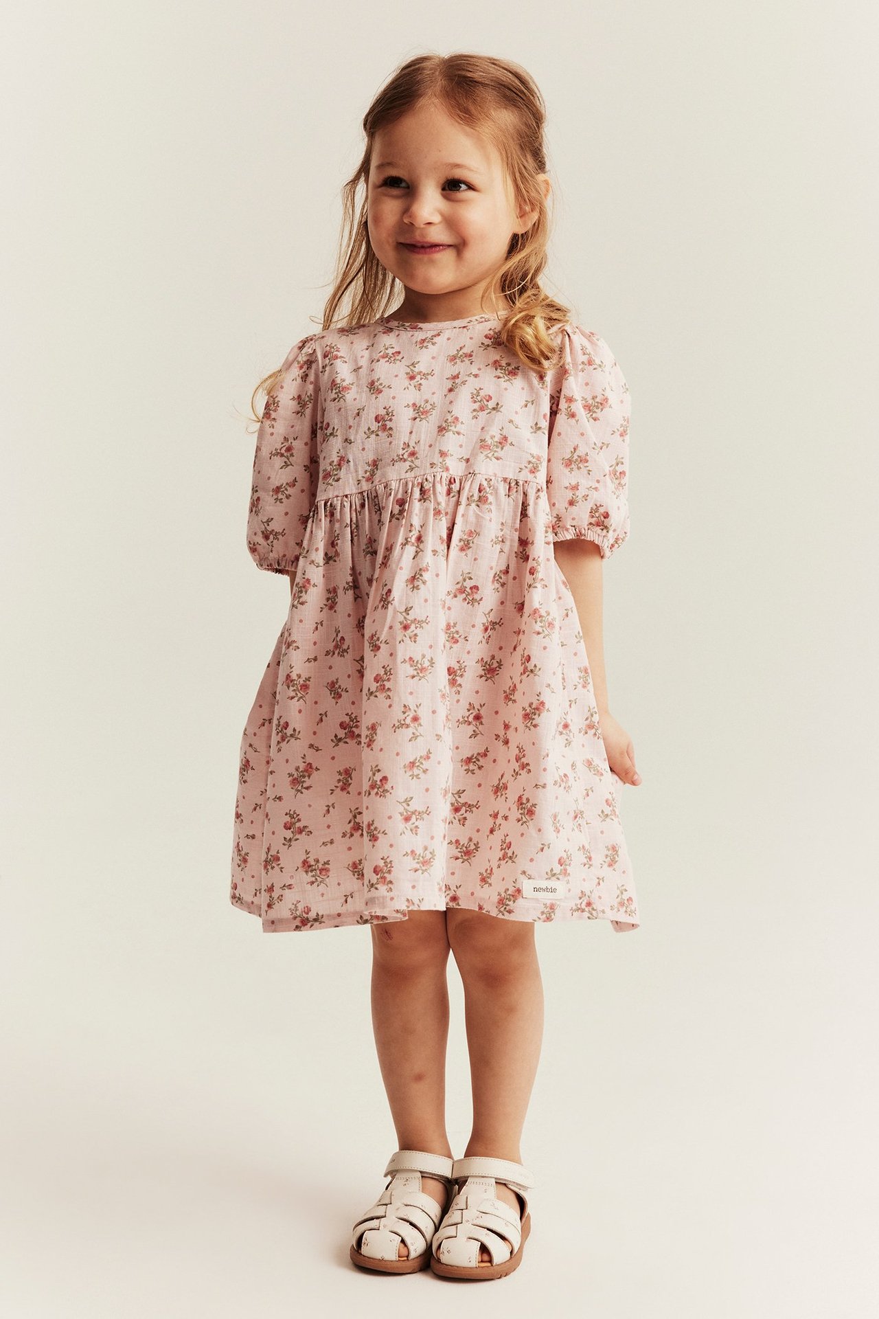 Front view of a kid girl wearing a light pink floral puff-sleeved dress and light beige sandals.