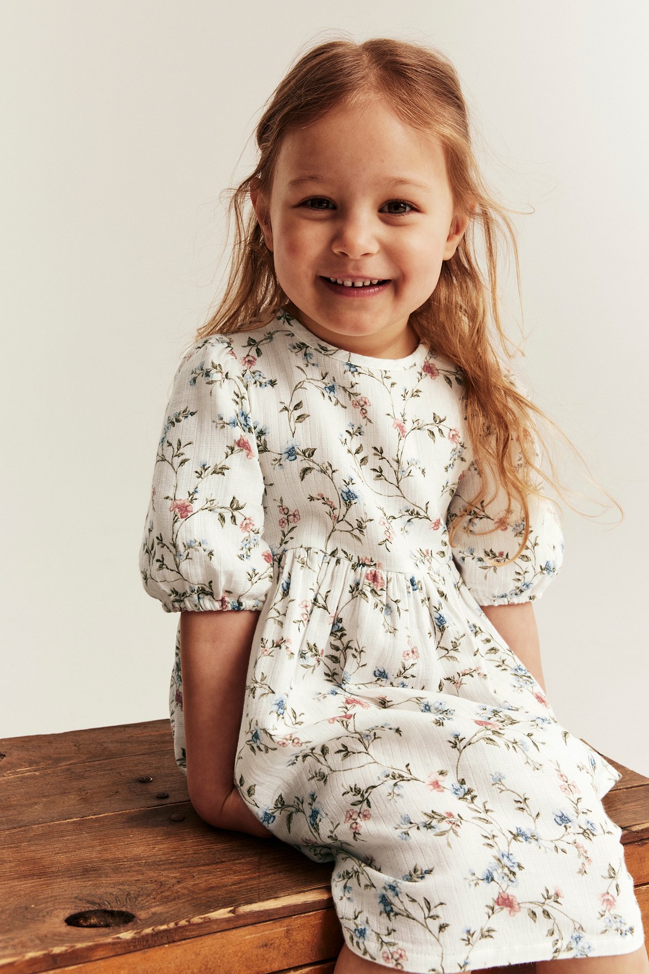 Front view of a smiling kid wearing an off-white floral dress with puff sleeves and elastic cuffs.