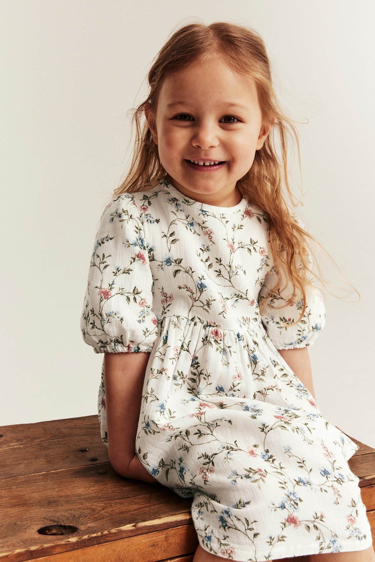 Front view of a smiling kid wearing an off-white floral dress with puff sleeves and elastic cuffs.