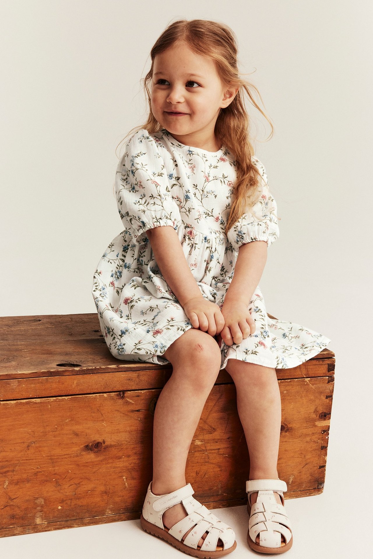 Front-side view of a kid in an off-white floral puff-sleeved dress and light sandals.