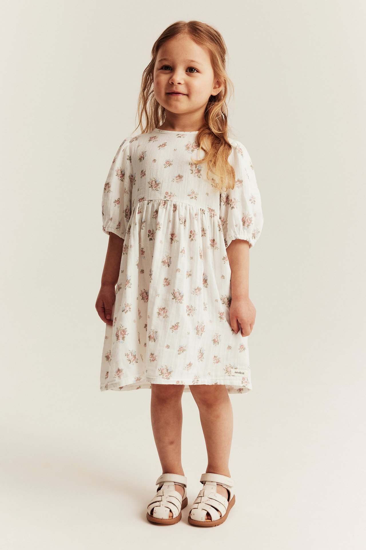 Front view of a kid wearing an off-white floral puff-sleeve dress and white sandals.