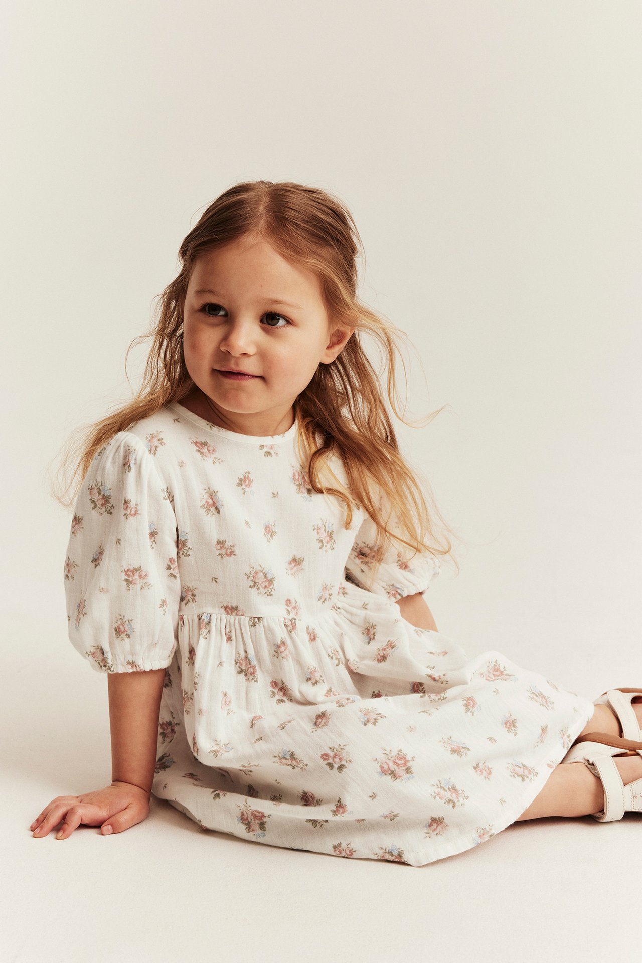 Frontansicht eines Kindes in einem cremeweißen Baumwollkleid mit Blumenmuster, Puffärmeln und weißen Sandalen.