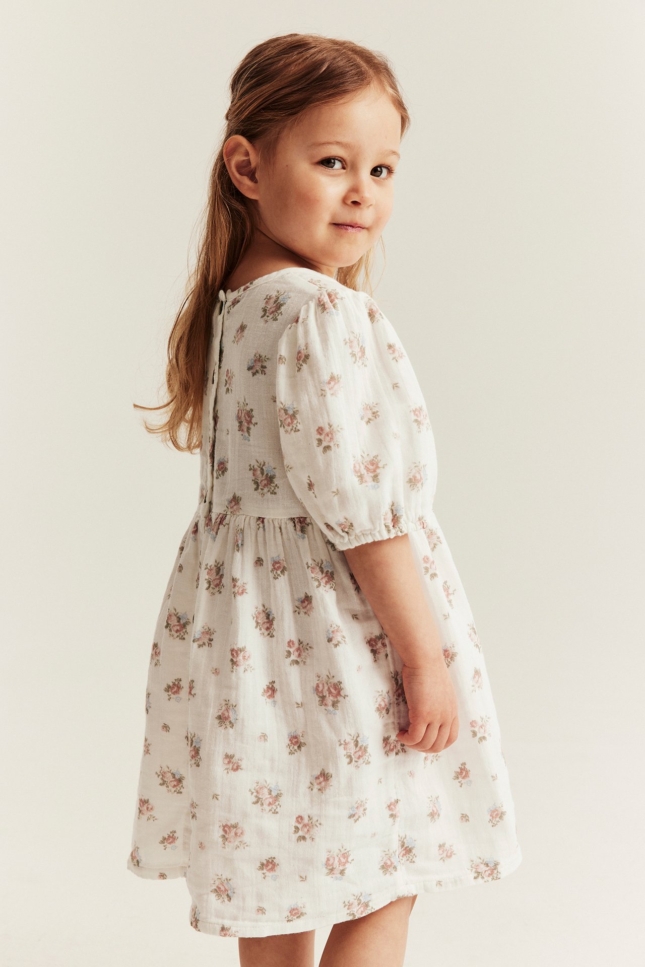 Side-back view of a kid wearing an off-white floral dress with puff sleeves and back buttons.