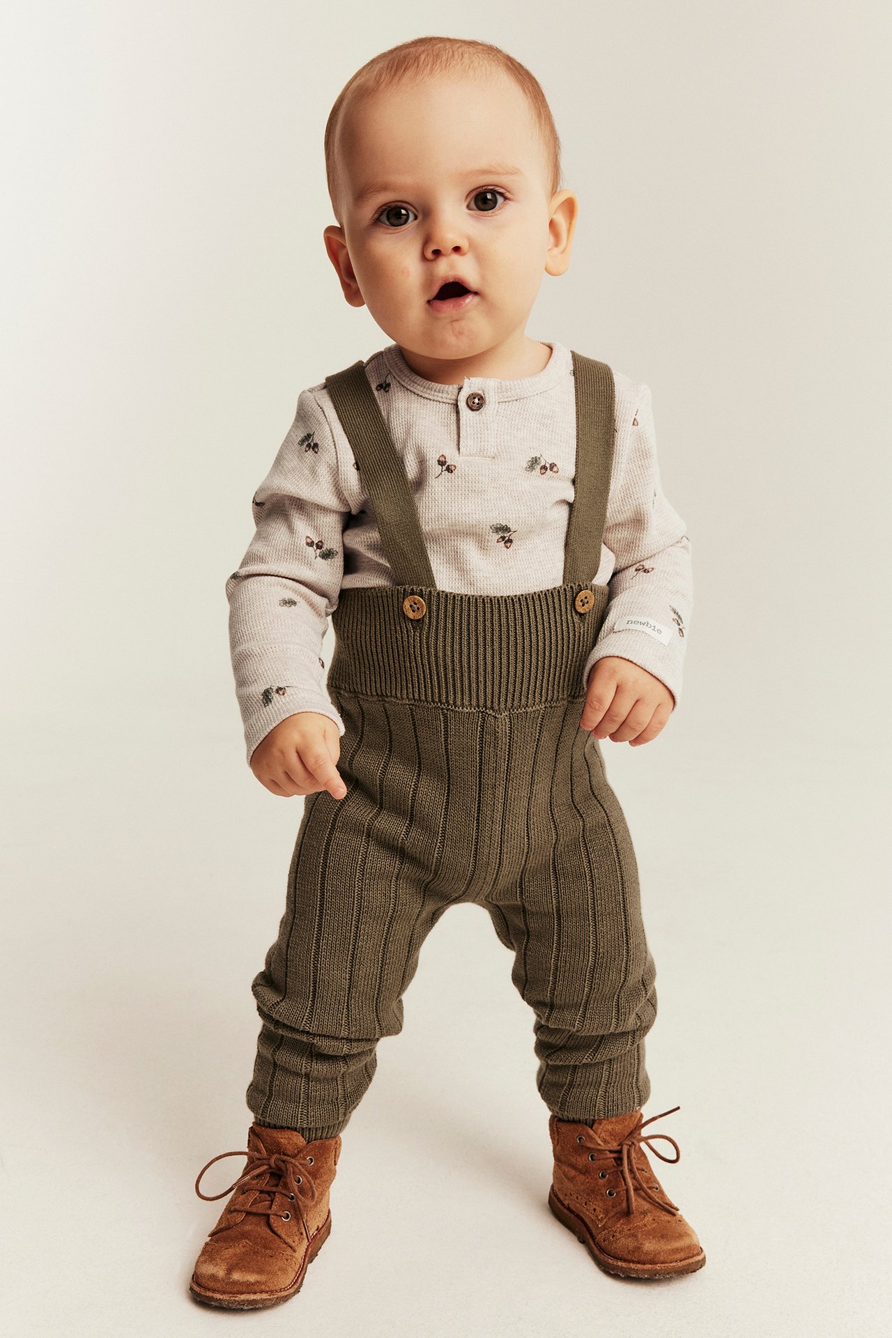 Vorderansicht eines Babys in olivgrünem Strick-Overall, beigem gemustertem Langarmshirt und braunen Schnürstiefeln.