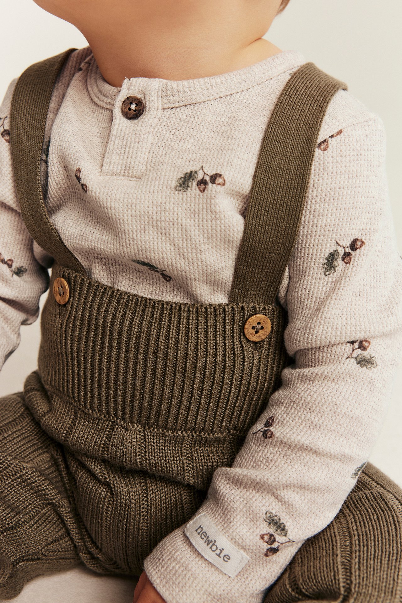 Nahaufnahme eines Babys in grünem Stricklatzoverall und hellbeigem Waffelhenley mit Eicheldruck.