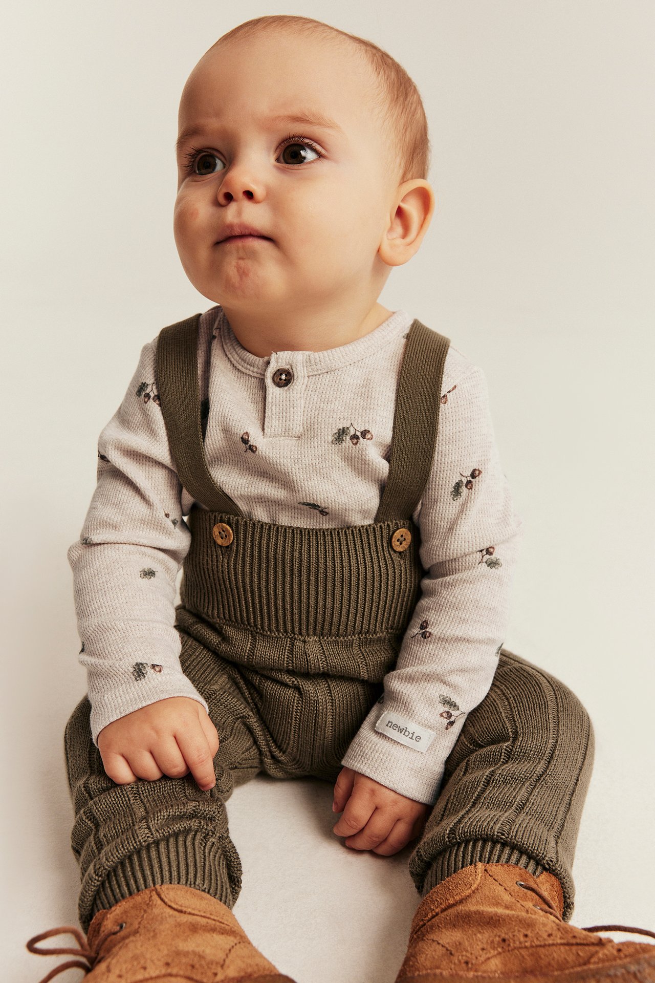 Baby, Vorderansicht: Olivgrüne gerippte Latzhose, Langarmshirt mit Eichel-Print, braune Stiefel.