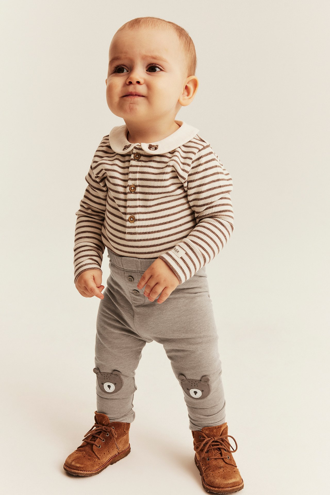 Front view of a baby wearing a brown striped top, gray bear-patch leggings, and brown boots.