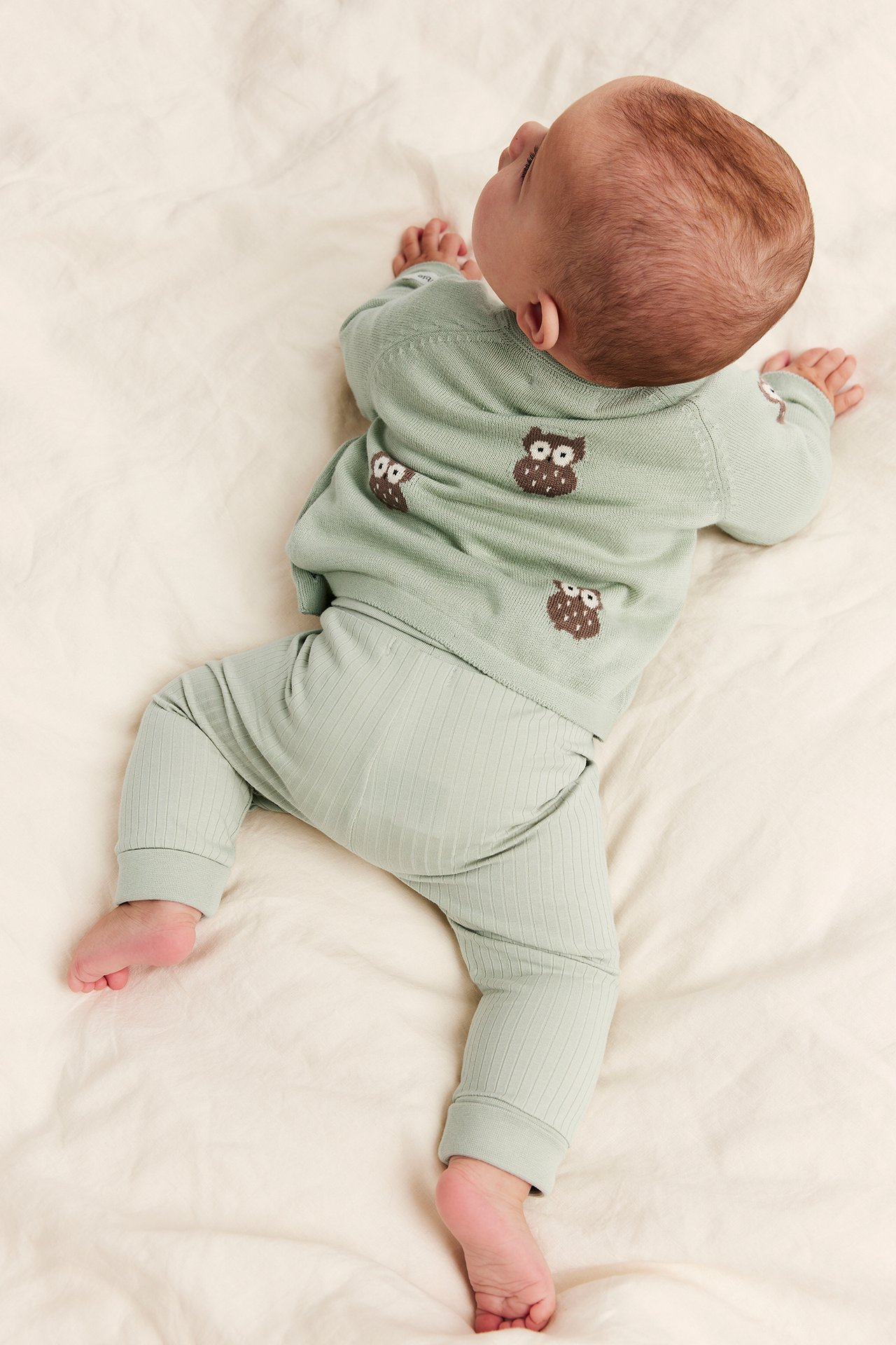 Back view of baby in a light green knit owl print sweater and matching ribbed leggings.
