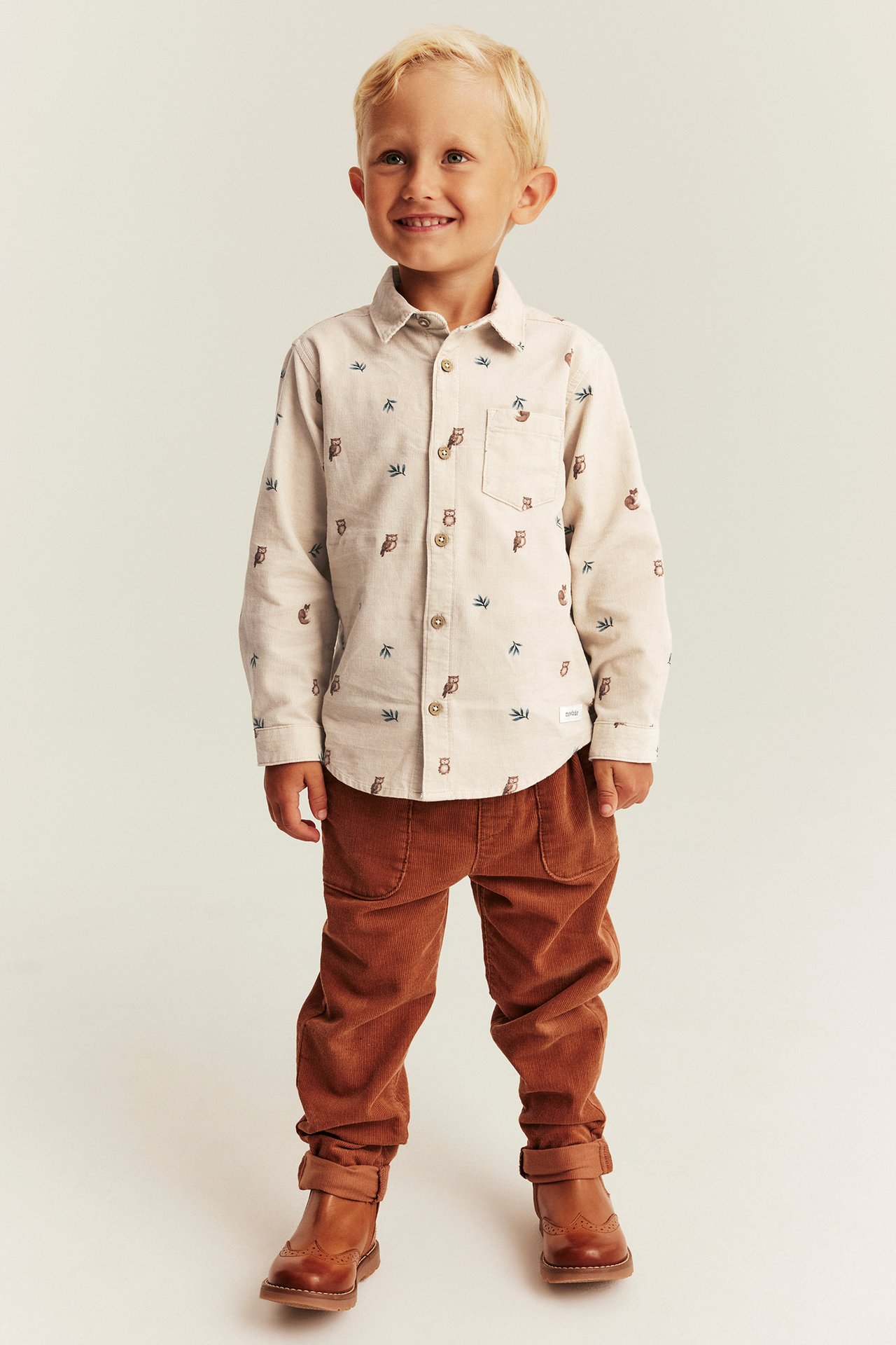Front view of a kid wearing a beige owl-patterned long-sleeved shirt, brown corduroy pants, and brown boots.
