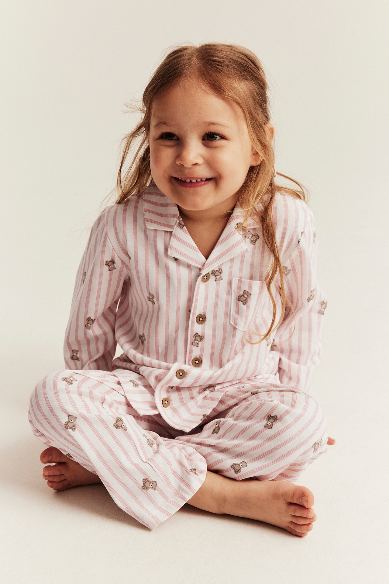 Front view: Kid wears light pink striped cotton pyjamas with teddy bears and wooden buttons.