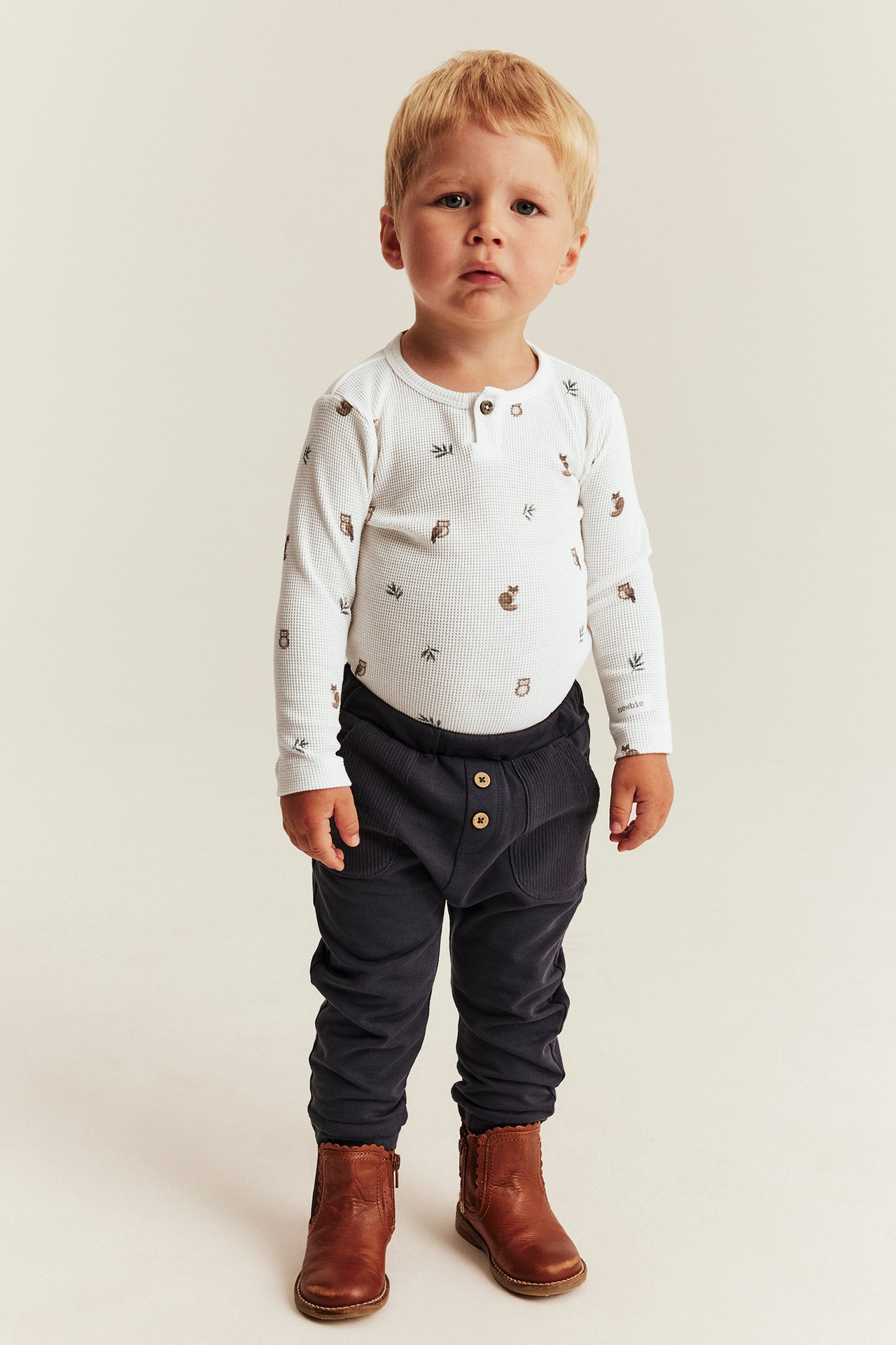 Front view of a toddler wearing a white long-sleeve animal print bodysuit, dark blue pants, and brown boots.