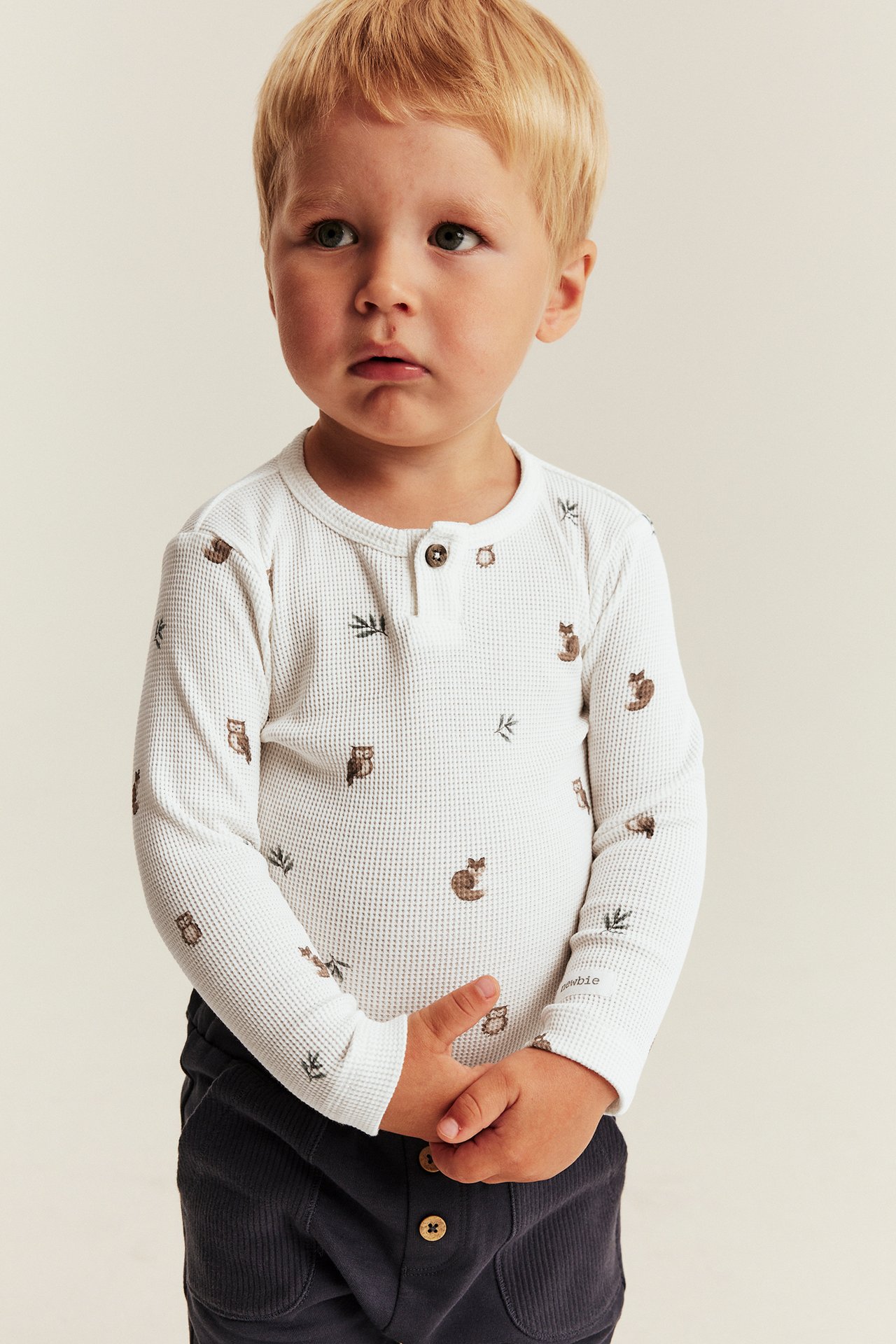 Front view of a kid wearing a white waffle knit long-sleeve top with animal print and dark gray corduroy pants.