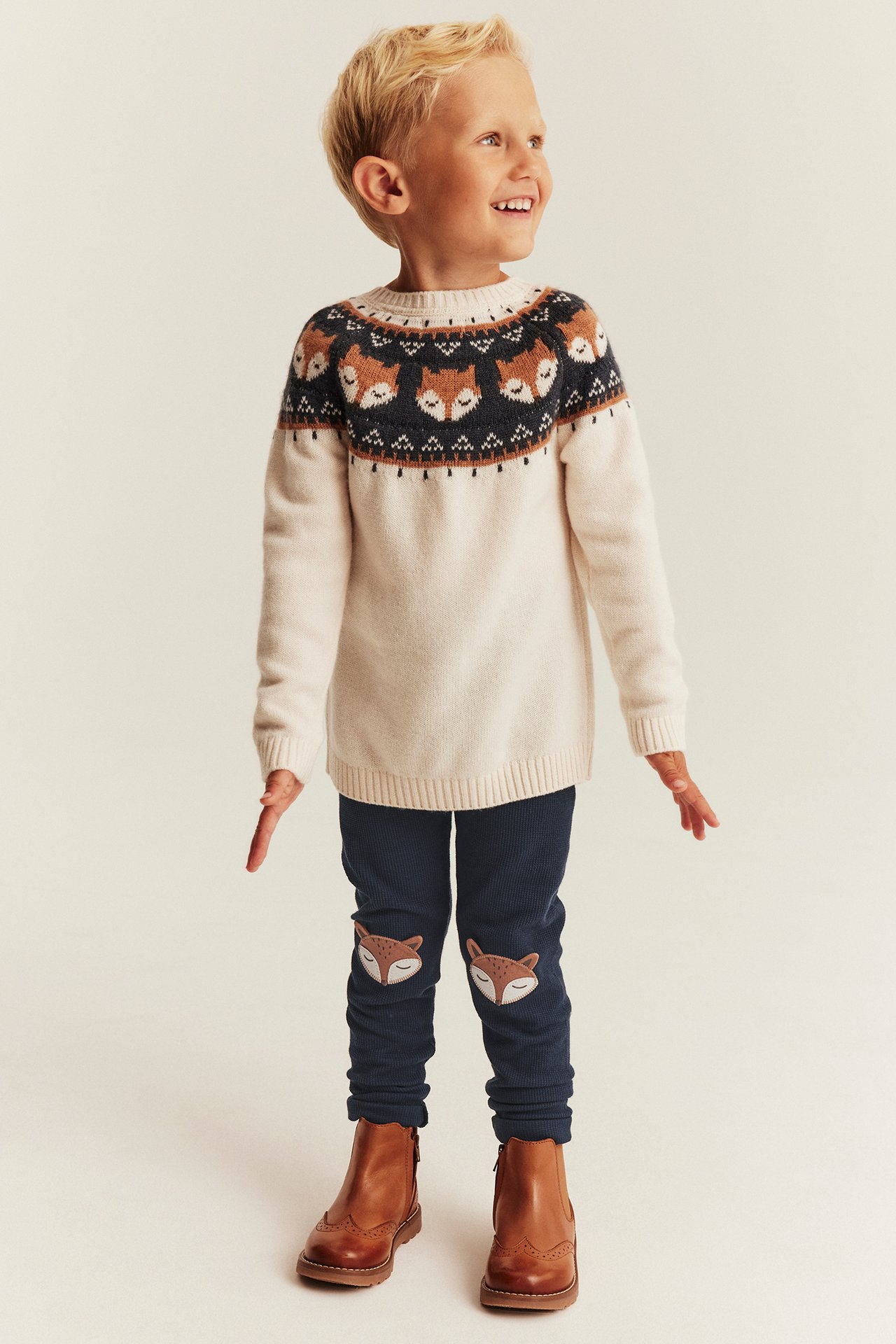 Vorn: Junge in cremefarbenem Fuchspullover, blauer Cordhose mit Fuchsflicken, braune Stiefel.