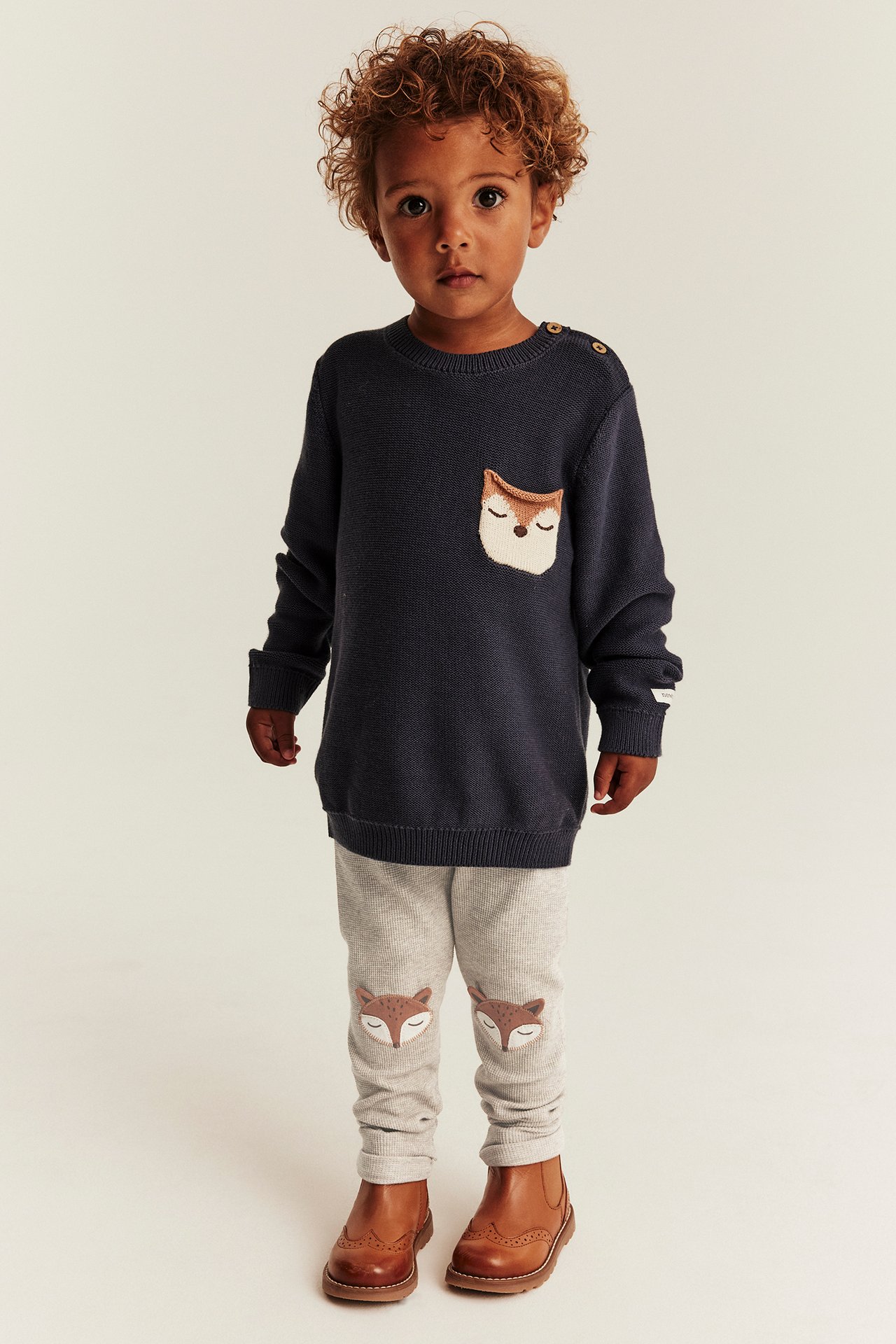 Vorderansicht eines Kindes in dunkelblauem Fuchs-Pullover, hellgrauen Fuchs-Leggings und braunen Stiefeln.