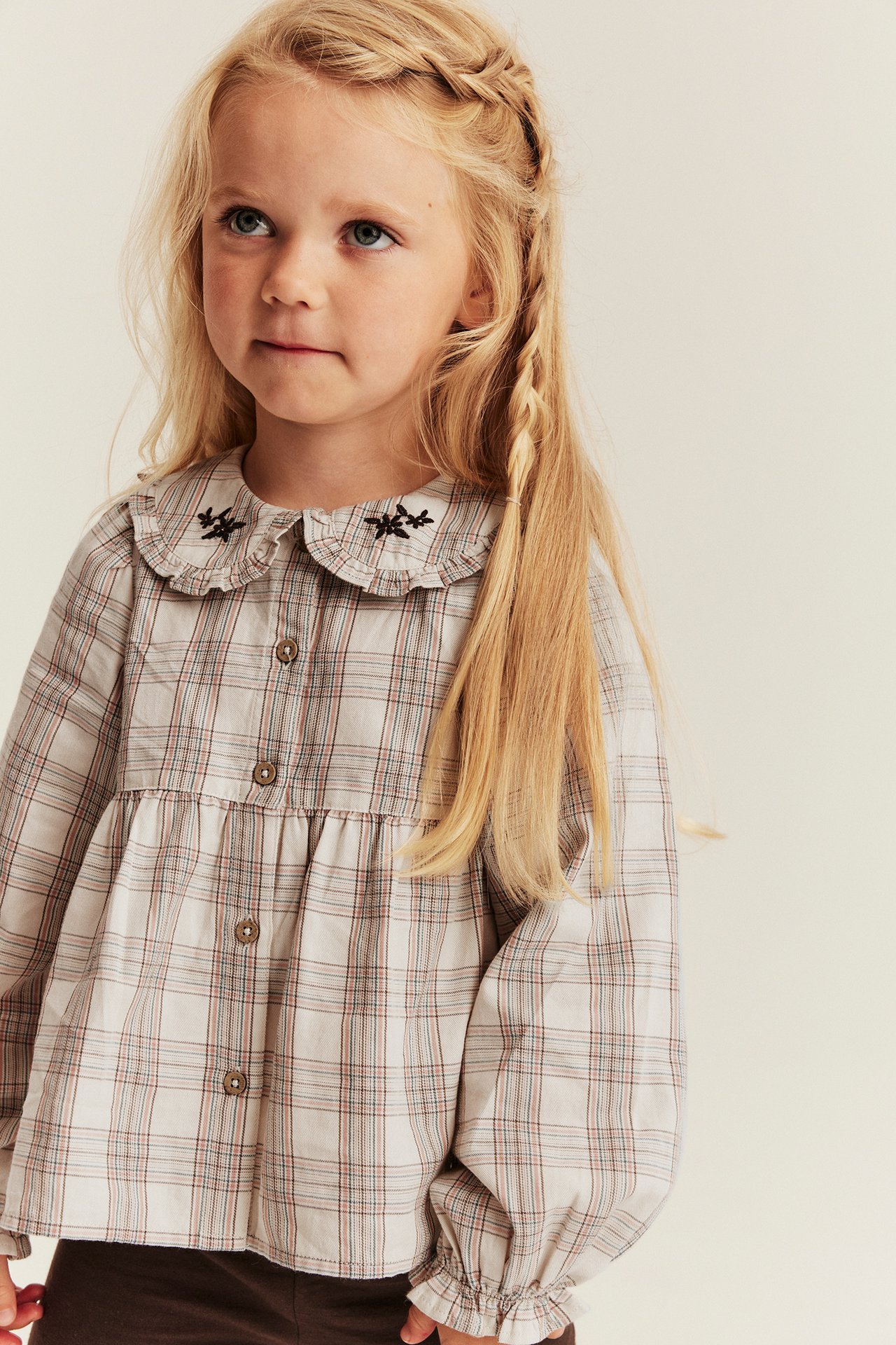 Front view of a kid's plaid long-sleeved shirt with a ruffled embroidered collar and brown pants.
