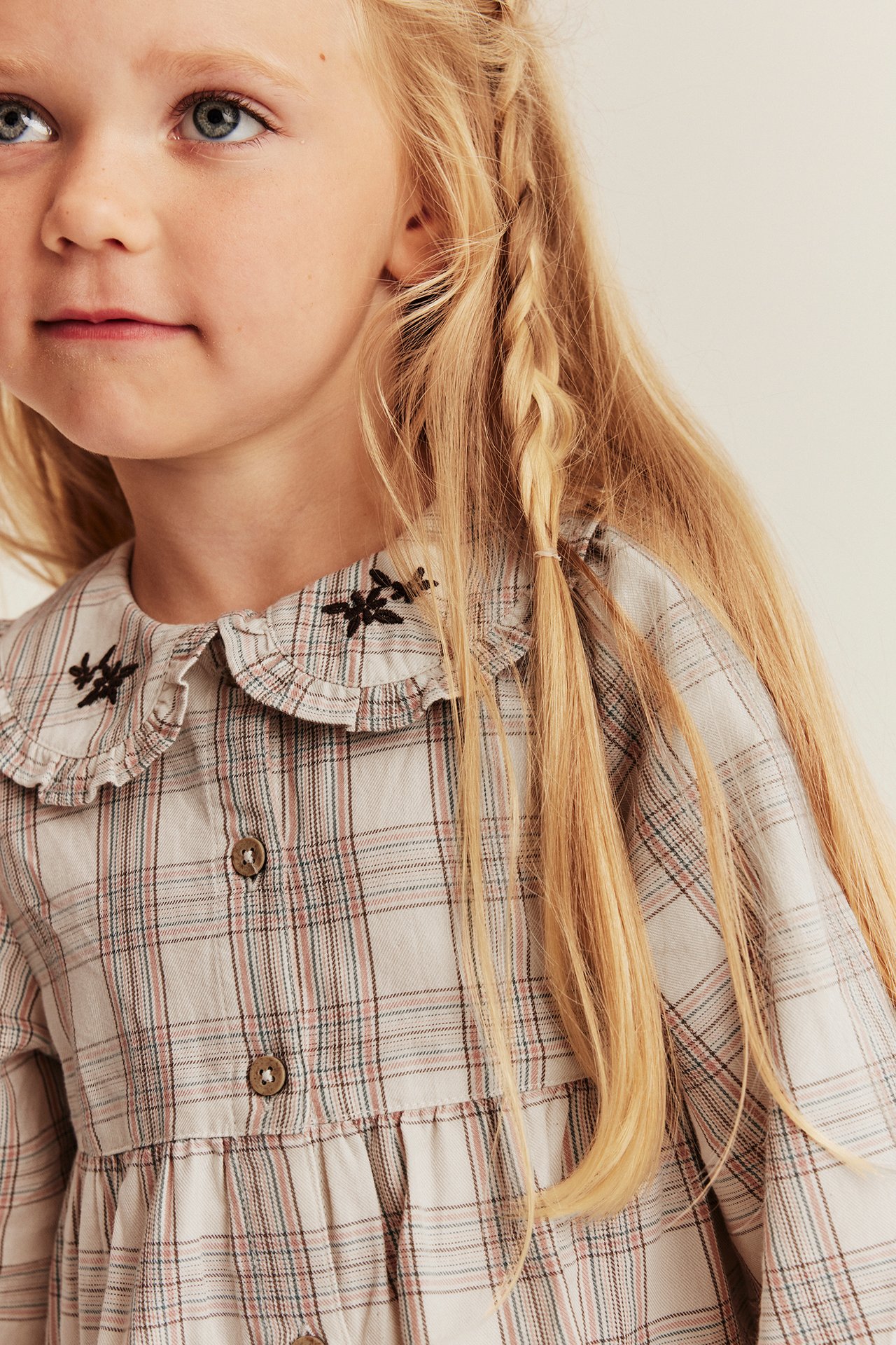 Close-up of a kid's beige plaid dress with a ruffled collar, floral embroidery, and front buttons.