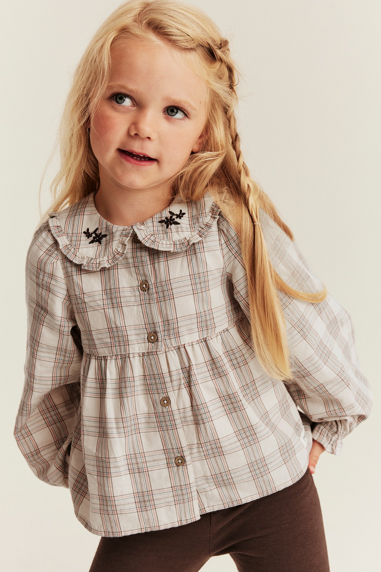 Front view of a kid wearing a beige plaid long-sleeve blouse with embroidered collar and brown leggings.