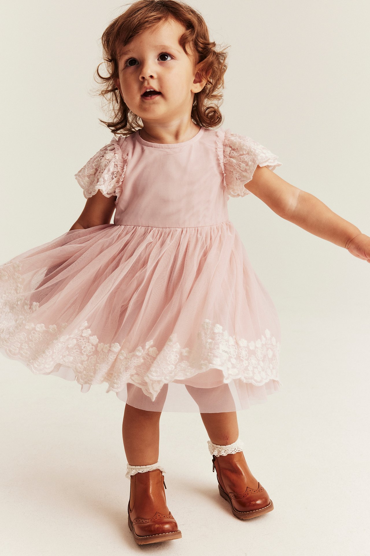 Baby in rosa kurzärmeligem Spitzen-Tüllkleid, rosa Leggings, weißen Rüschensocken und braunen Stiefeln.