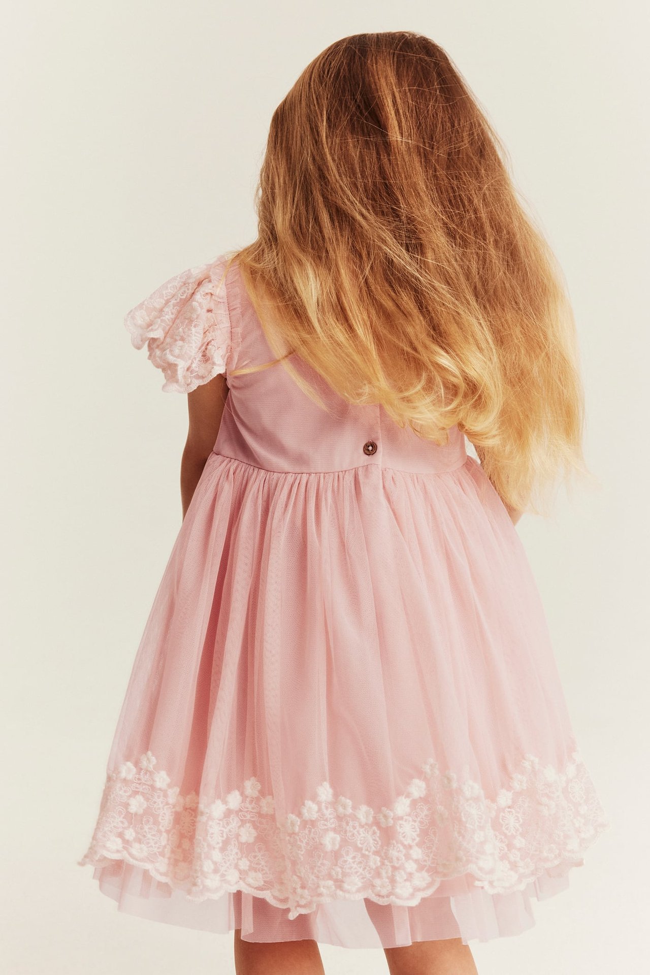 Back view of a kid wearing a pink tulle dress with lace sleeves, button, and decorative hem.