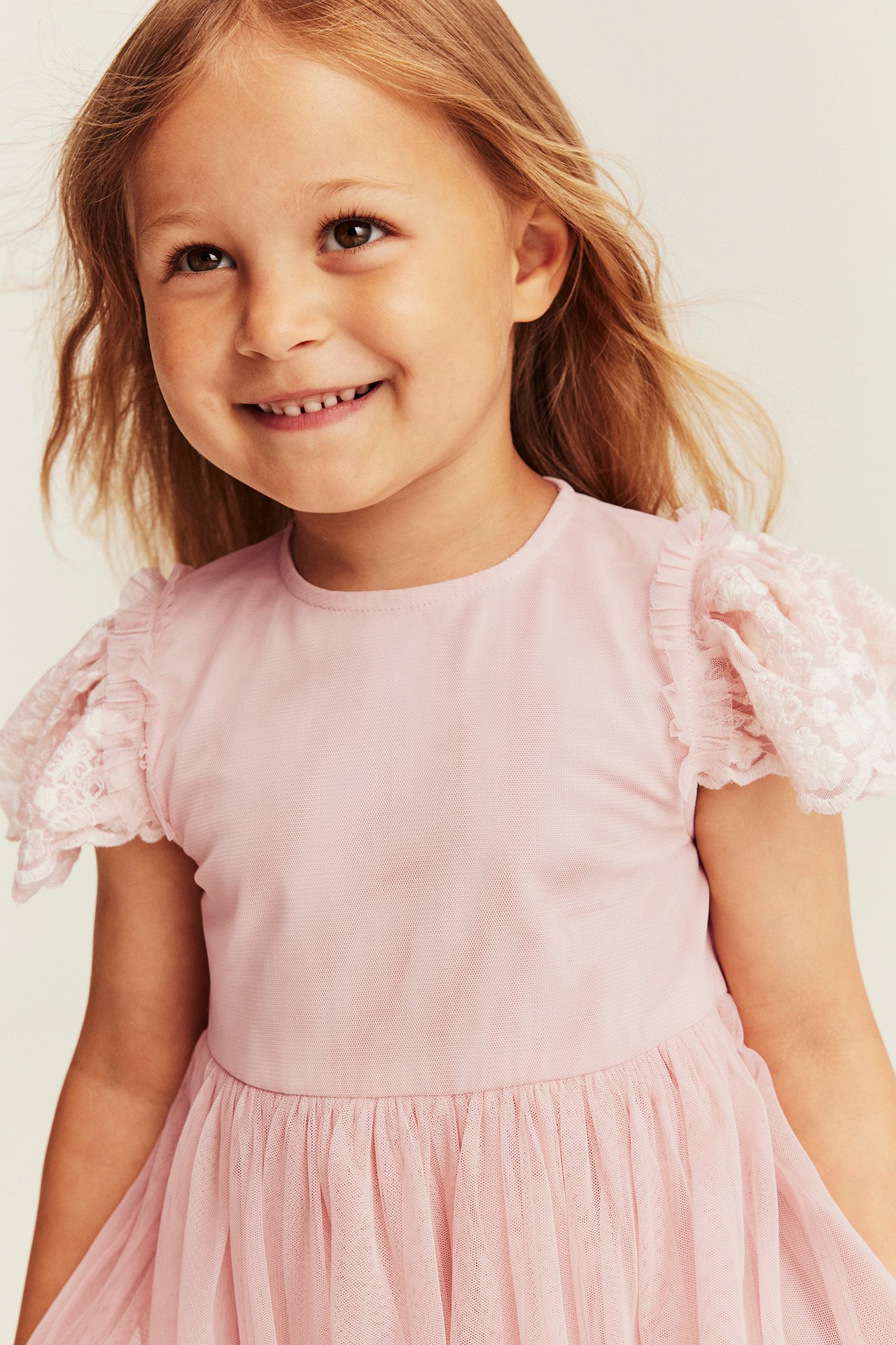 Front view of a smiling kid wearing a pink dress with ruffled lace puff sleeves and a tulle skirt.