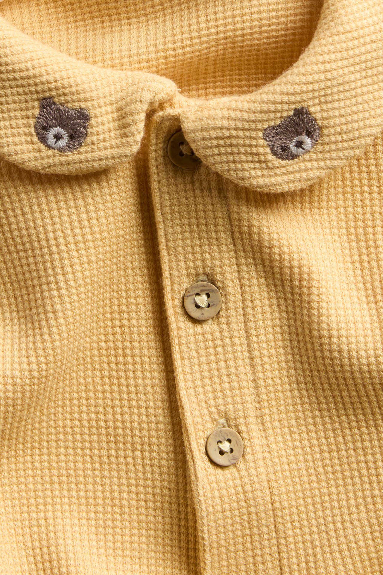 Close-up of a yellow waffle-knit baby top with embroidered bear heads on collar and buttons.