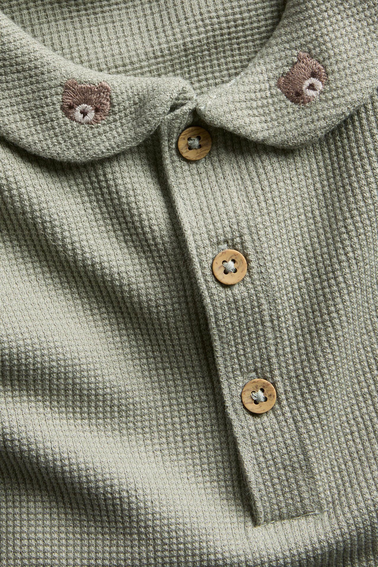 Close-up of a baby's sage green waffle knit top with embroidered bears on collar and wooden buttons.
