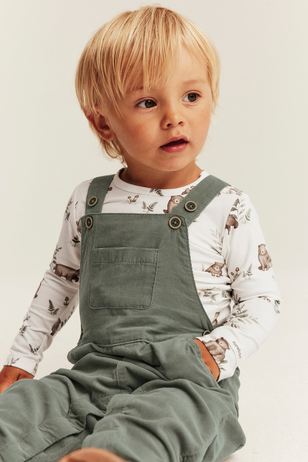 Front view of a kid wearing green corduroy overalls over a white bear-print long-sleeve top.