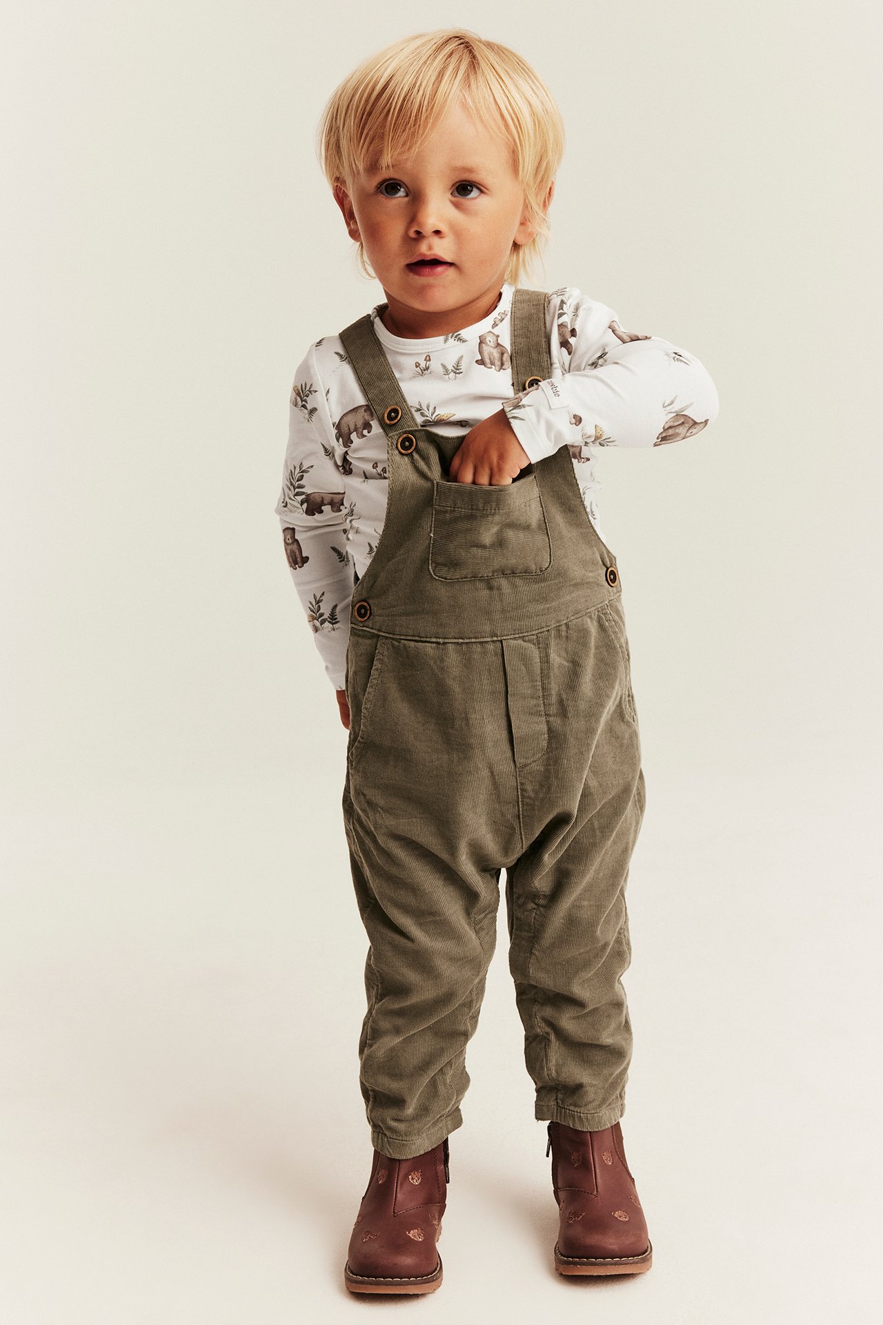 Kind in olivgrünem Cord-Overall, weißem Bären-Langarmshirt und braunen Stiefeln, Vorderansicht.