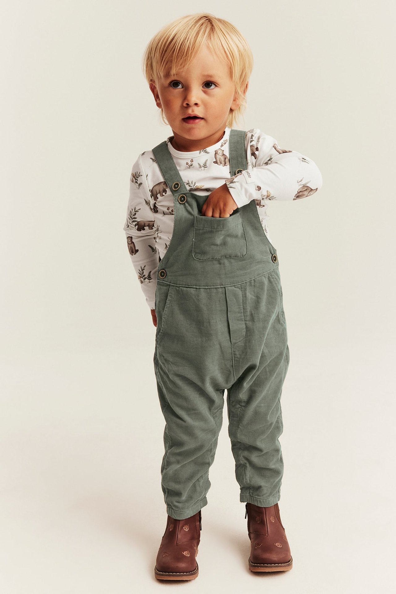Front view of a kid in sage green overalls over a white bear-print top, with brown boots.