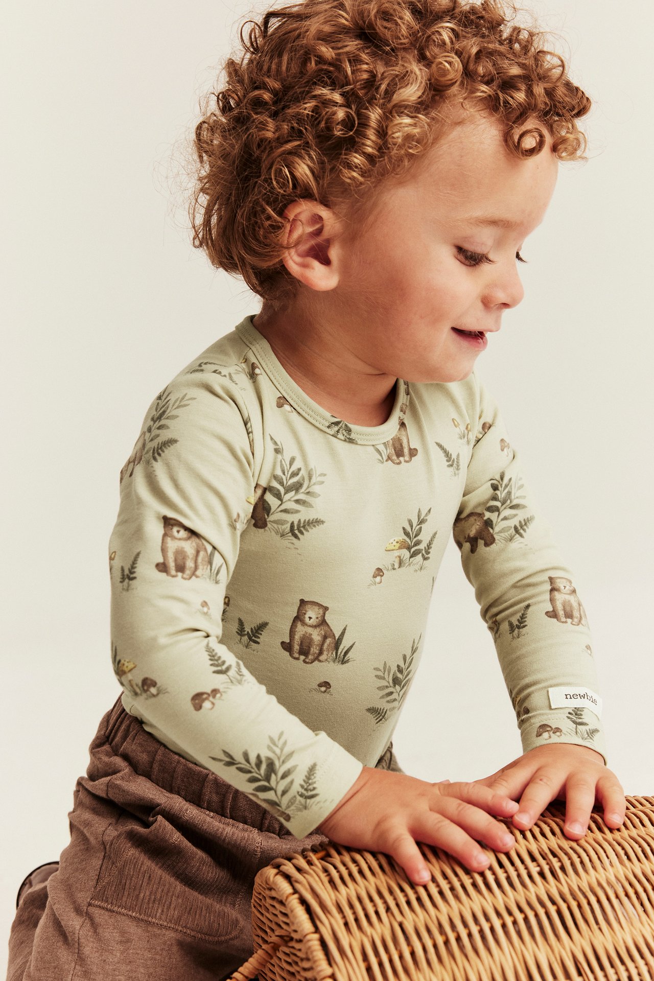 Kind in einem hellgrünen Langarmshirt mit Bärenmuster und braunen Rippenhosen, an einem Weidenkorb lehnend.