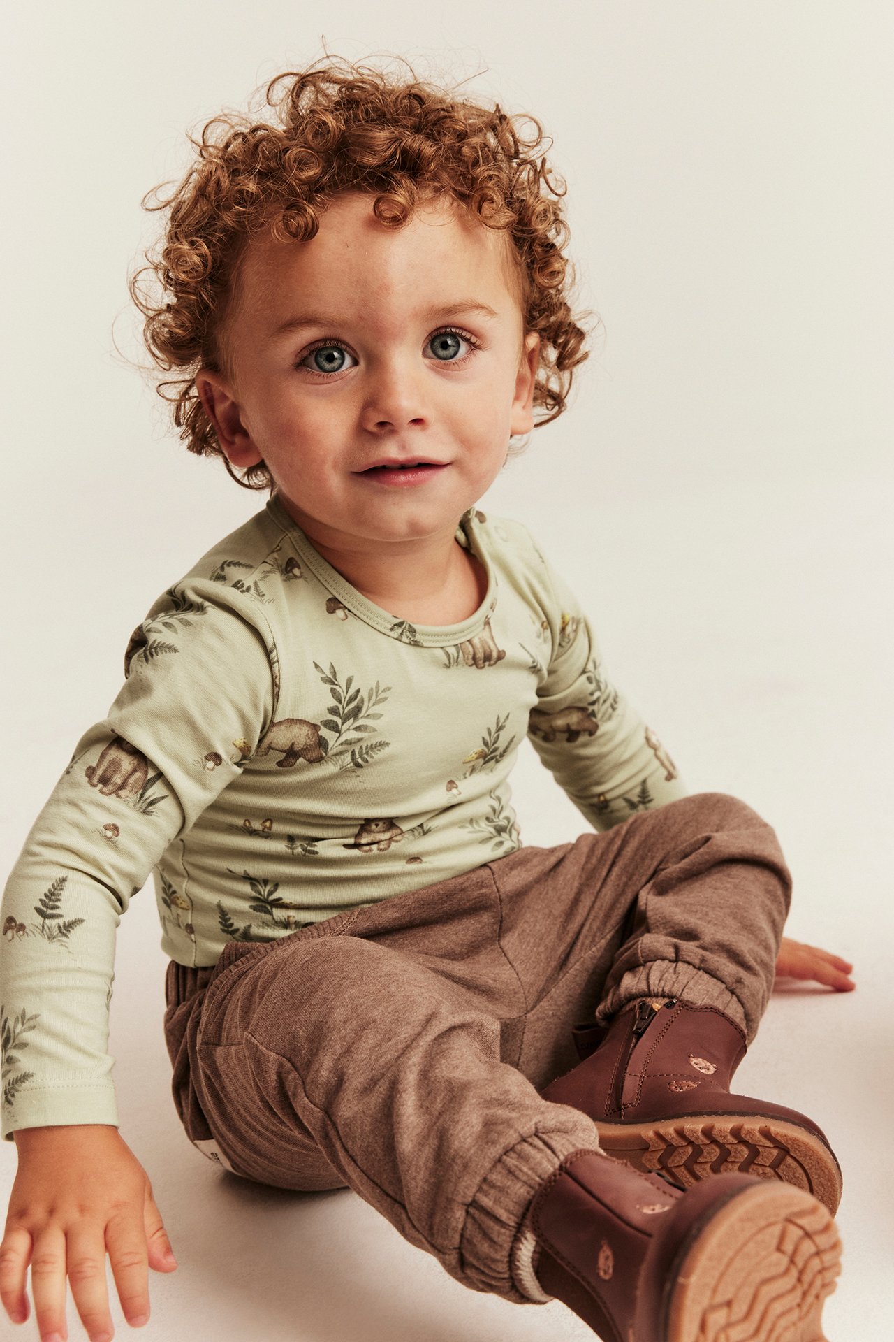 Vorderansicht Baby in hellgrünem Langarmshirt mit Waldtierdruck, brauner Hose und braunen Stiefeletten.