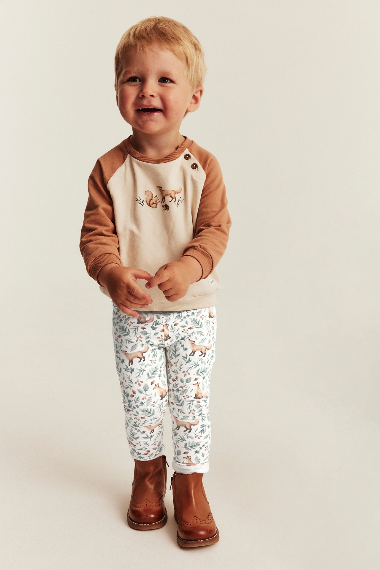 Vorderansicht: Kind in besticktem beige-braunen Langarmshirt, weißen gemusterten Leggings und braunen Stiefeln.