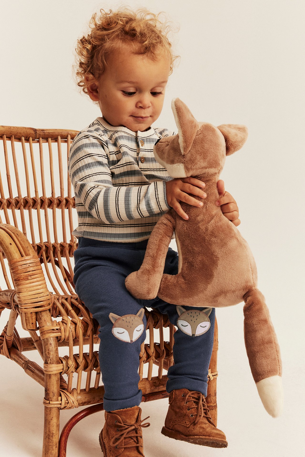 Front view of a baby in blue ribbed fox-patch leggings, striped top, brown boots, holding a fox toy.
