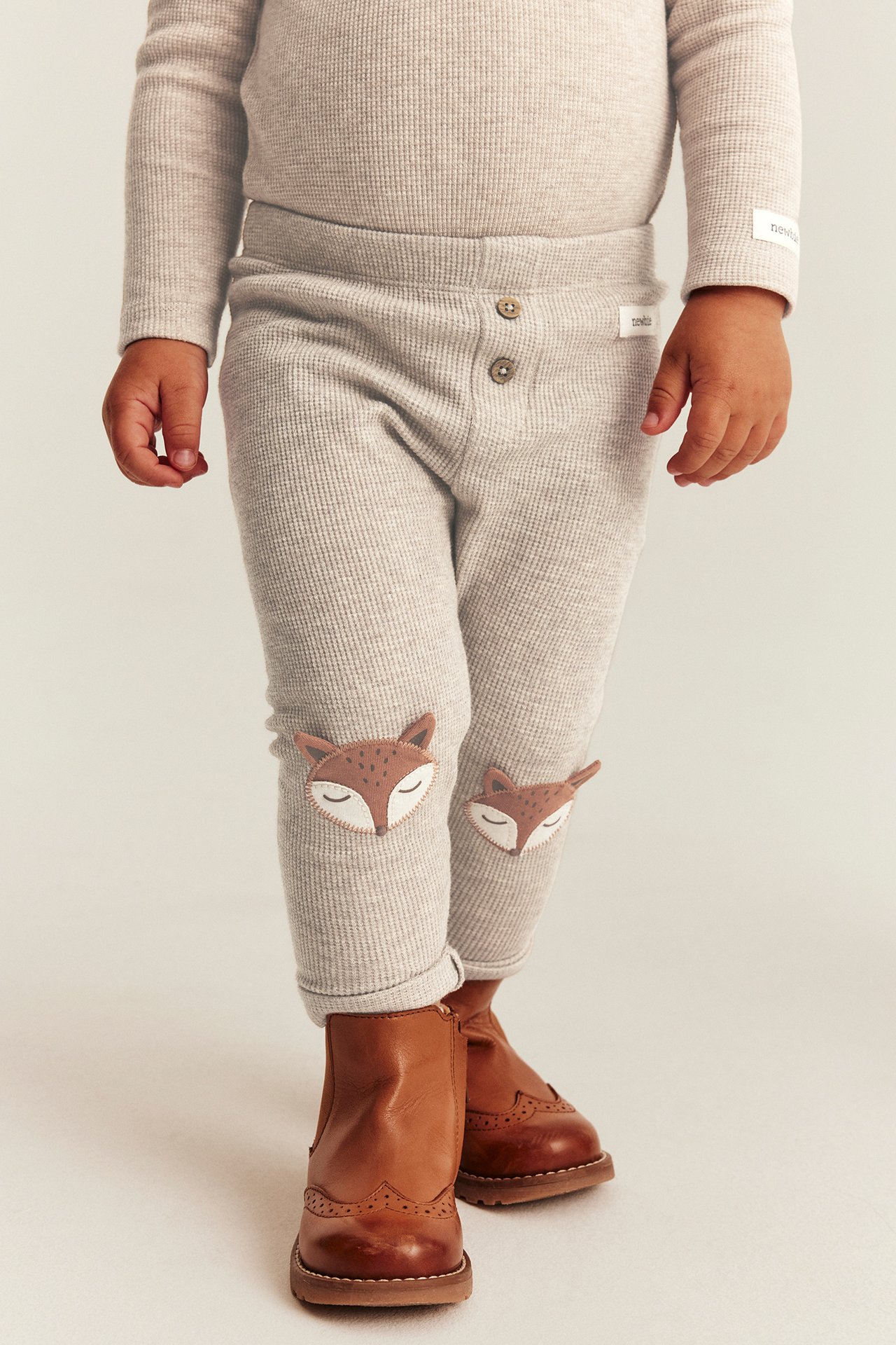 Vorderansicht: Baby in hellbeiger Ripphose mit Fuchsknie-Patches, passendem Oberteil, braunen Stiefeln.