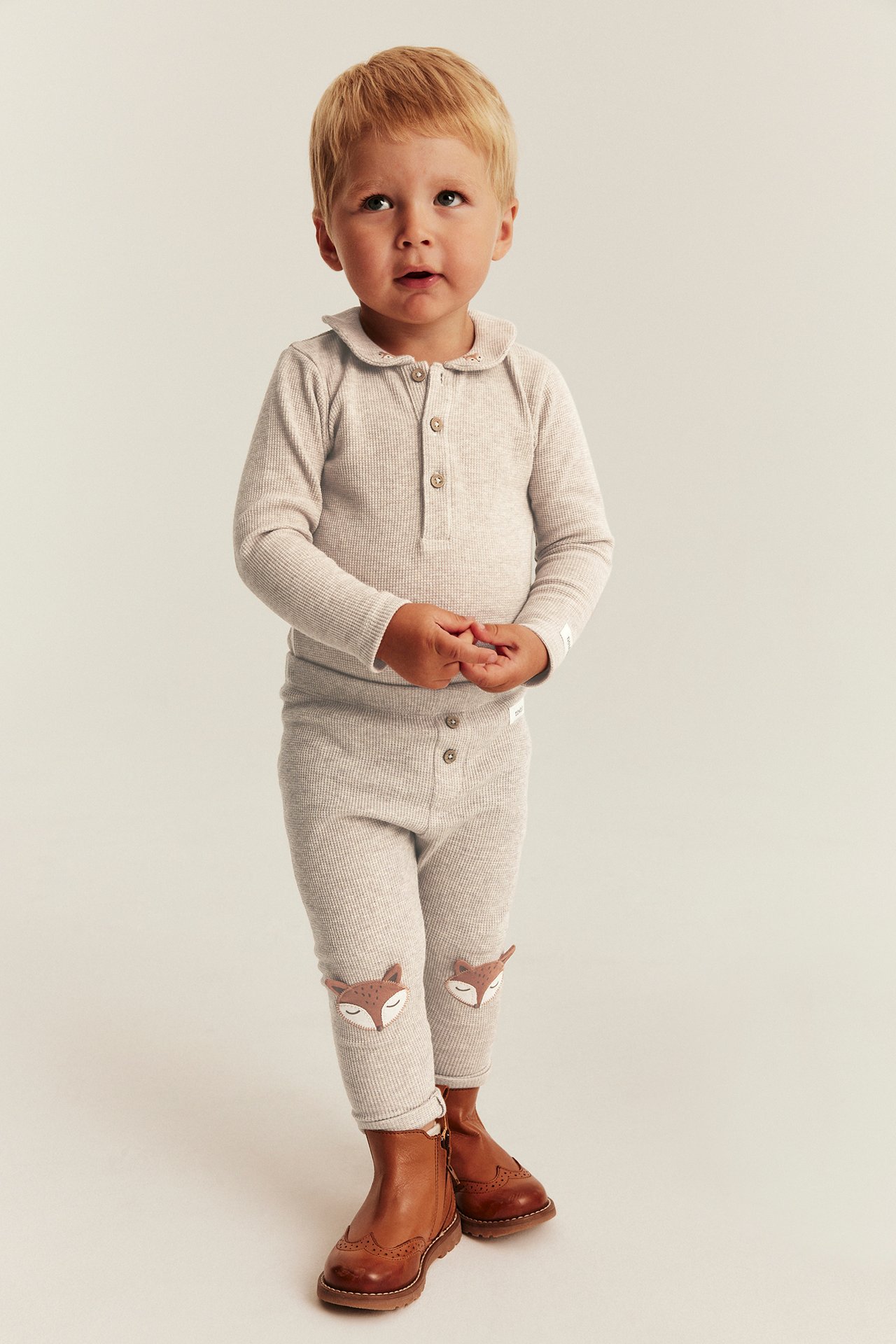 Frontansicht Baby in hellbeigem Rippstrick-Set mit Fuchs-Knie-Patches und braunen Stiefeln.