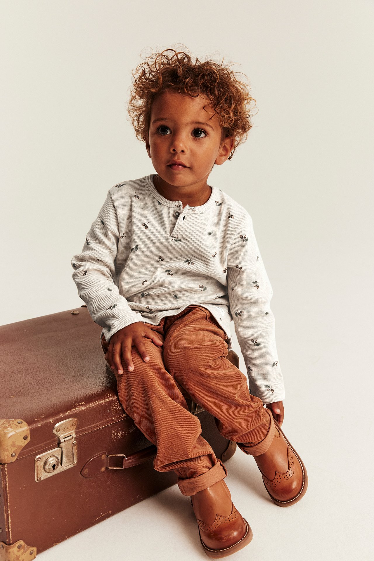 Kid in light gray printed henley shirt, brown corduroy pants, brown brogue shoes.