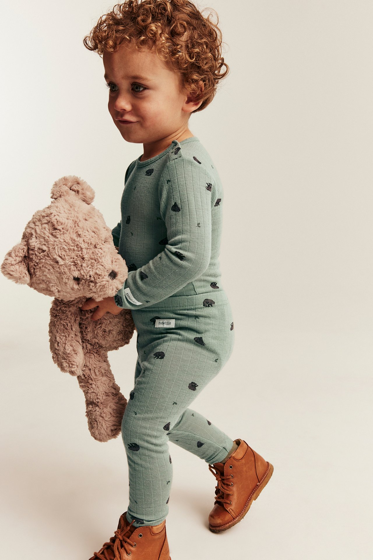 Side view of kid in sage green ribbed set with bear print, holding teddy, brown boots.